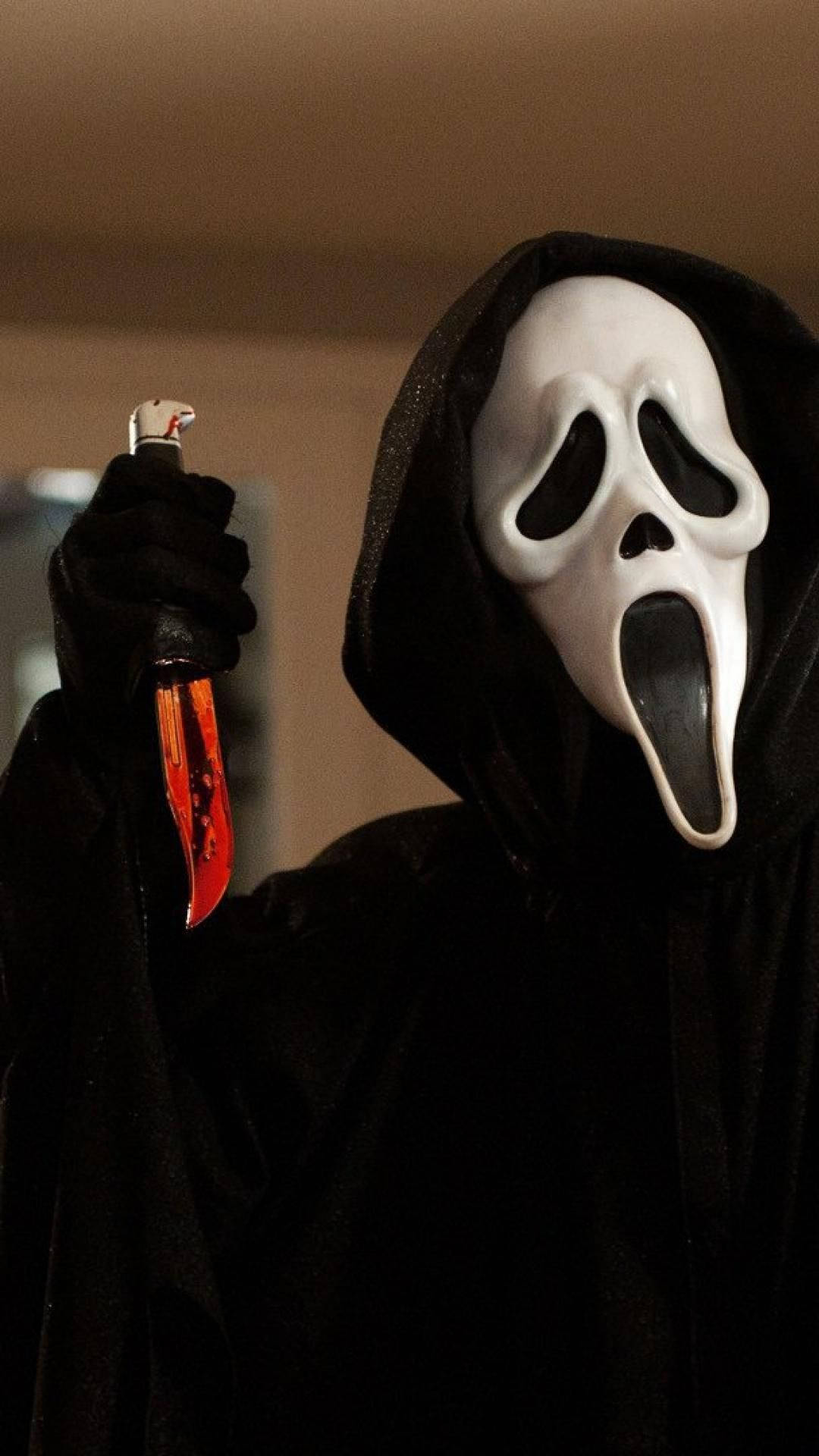 Scream 桌布 壁紙 — 一個戴著白色Ghostface面具、身穿黑色連帽長袍的人，在燈光昏暗的房間裡拿著一把沾滿血的刀。