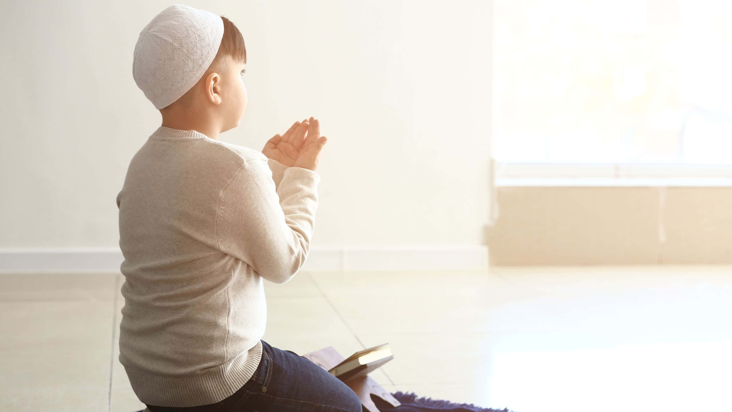 Praying 桌布 壁紙 — 一個戴著白色基帕的小男孩跪在室內的祈禱毯上，雙手合十祈禱，柔和的陽光從他身後的窗戶照進來。