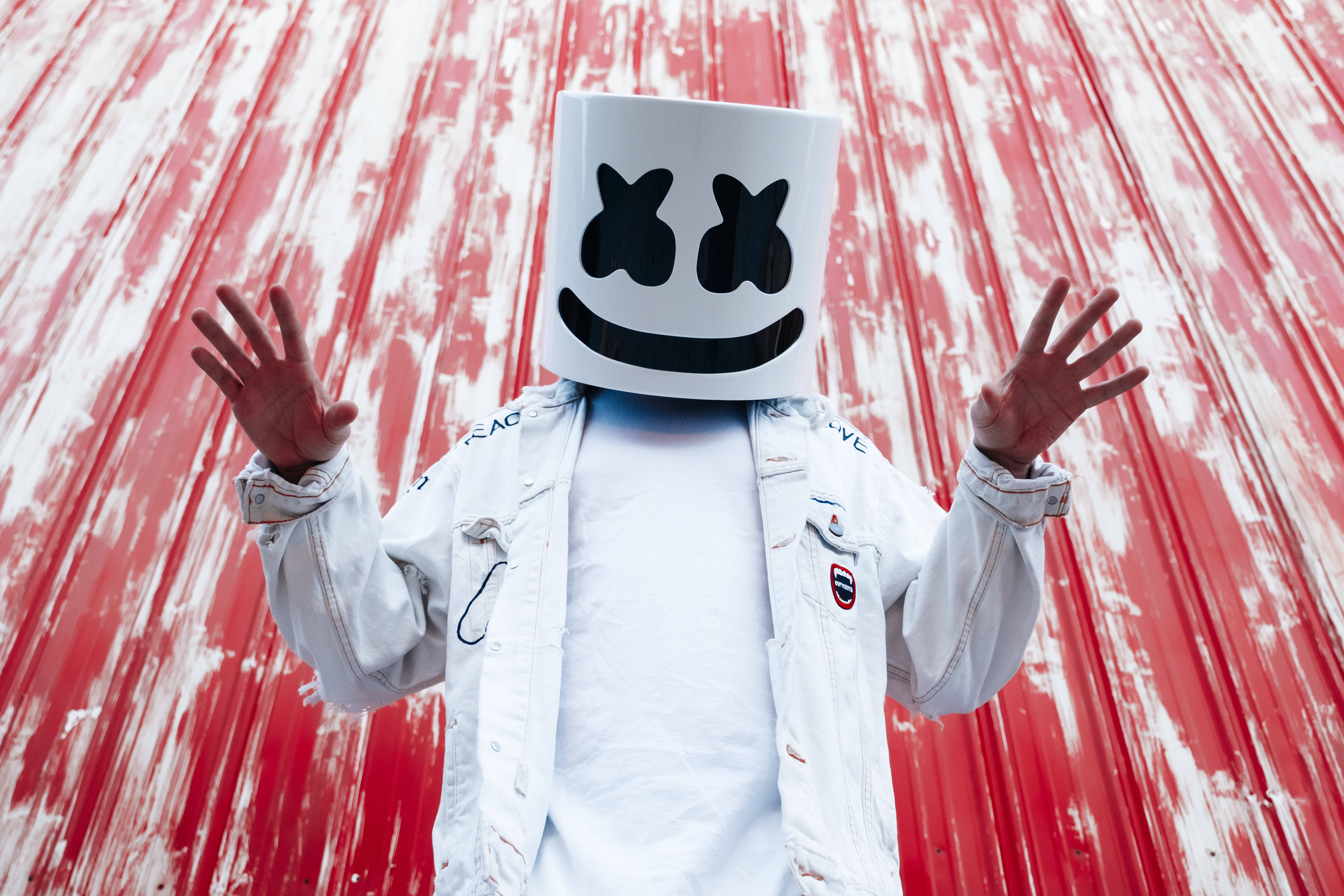 marshmello 4K 桌布 壁紙 — 一個戴著白色棉花糖頭盔和外套的人，舉起雙手，站在風化的紅白波紋牆前。
