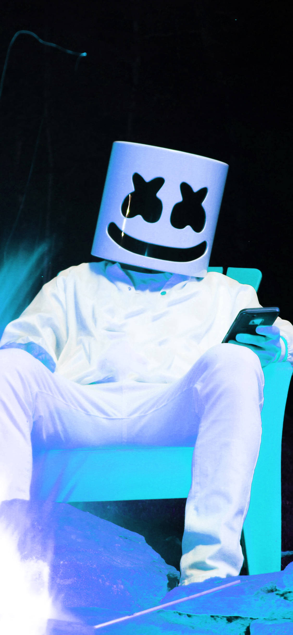 Marshmello 動態圖 桌布 壁紙 — 一個戴著白色棉花糖頭盔和穿著白色棉花糖服裝的人坐在發光的藍色椅子上，在舞台燈光下拿著電話。