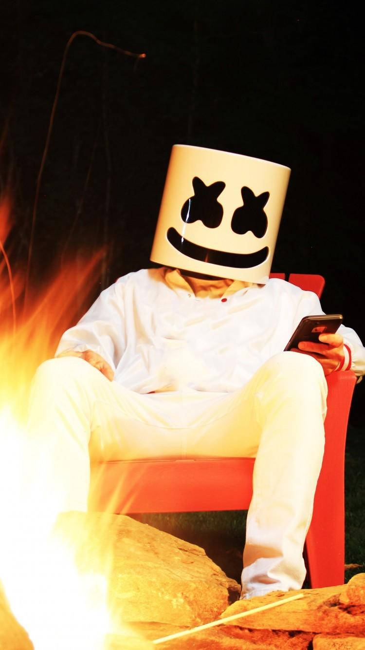 marshmello iPhone 桌布 壁紙 — 一個戴著白色棉花糖頭盔的人晚上坐在紅色椅子上，旁邊是熊熊燃燒的營火。