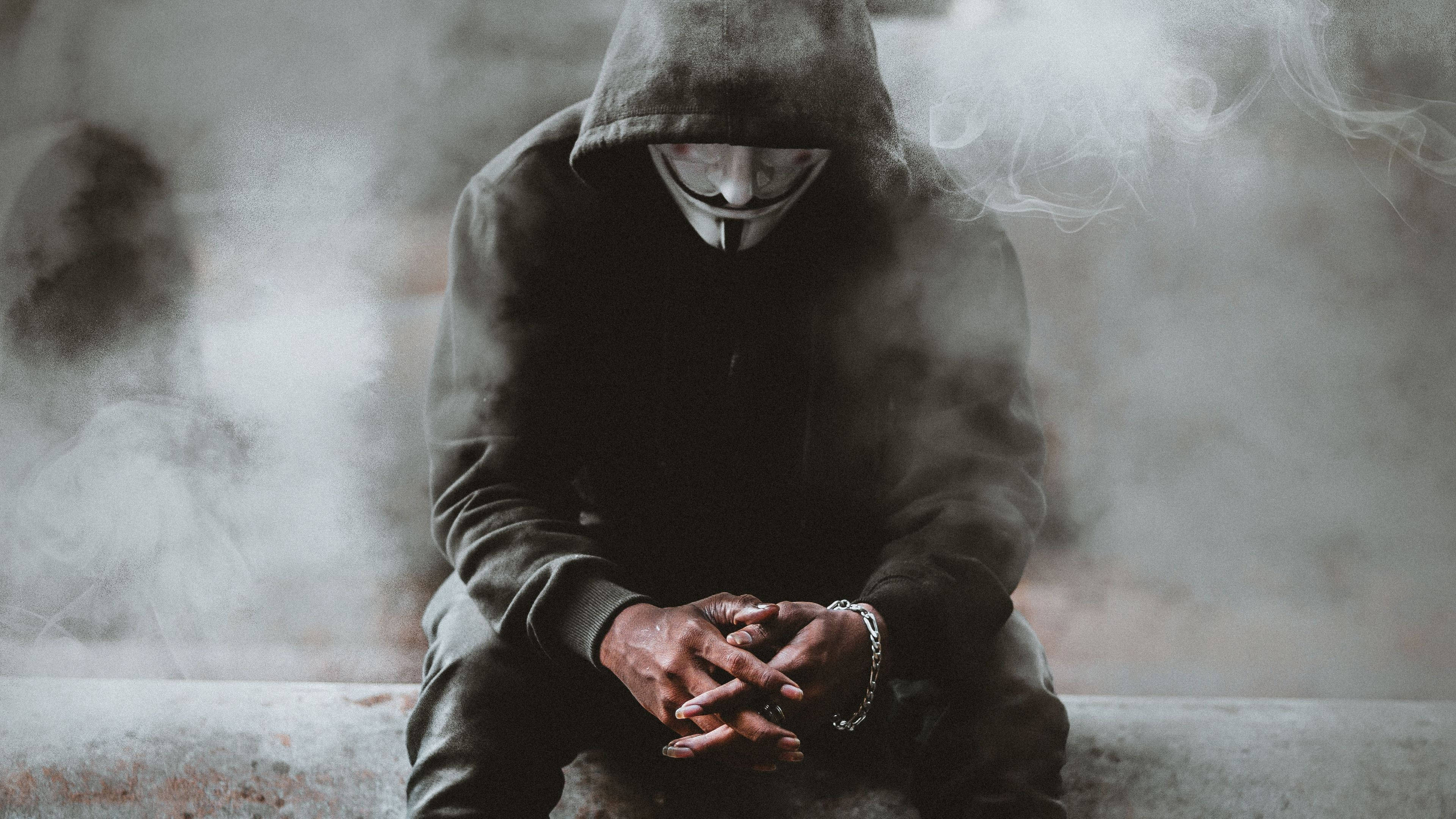 Hoodie 桌布 壁紙 — 一個戴著白色蓋伊·福克斯面具的兜帽人物，蜷縮在煙霧般的灰色調中，雙手緊握，可見一條鏈條手鐲。