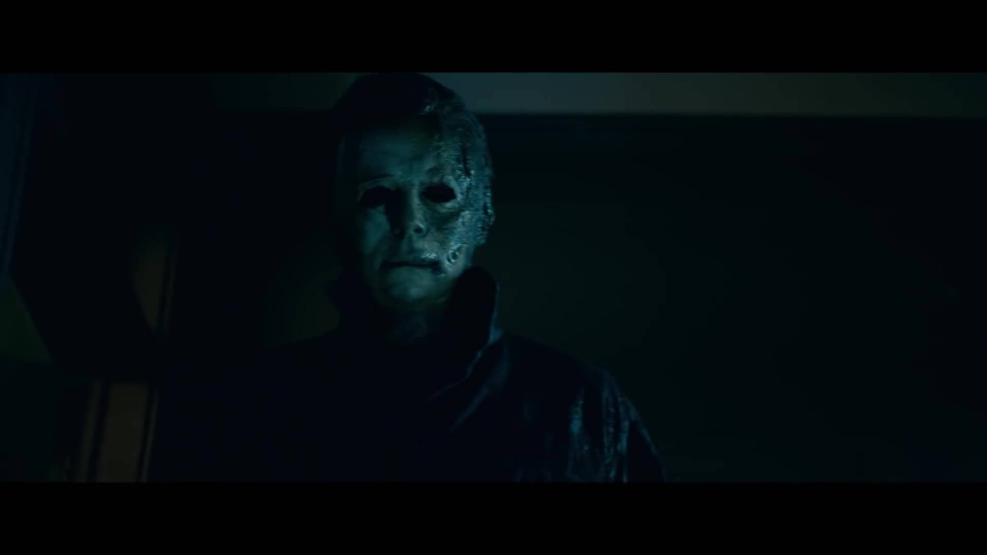 Halloween Kills 桌布 壁紙 — 一個戴著白色面具、身穿黑色外套的威脅人物，站在一個昏暗、帶有詭異藍光的房間裡。