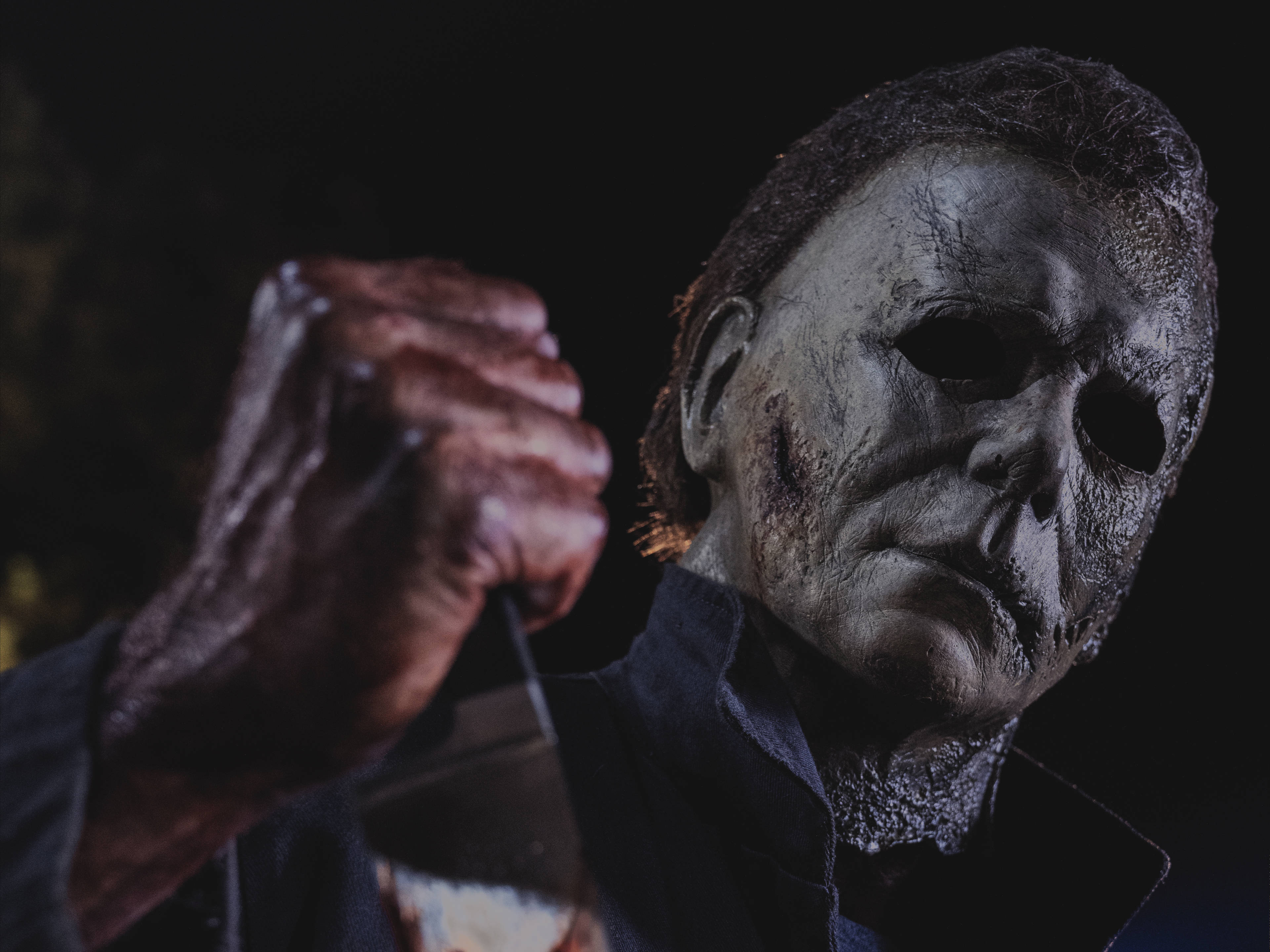 Halloween Kills 桌布 壁紙 — 一個戴著破舊白色面具的威脅人物，在黑暗背景下握著一把血淋淋的刀。