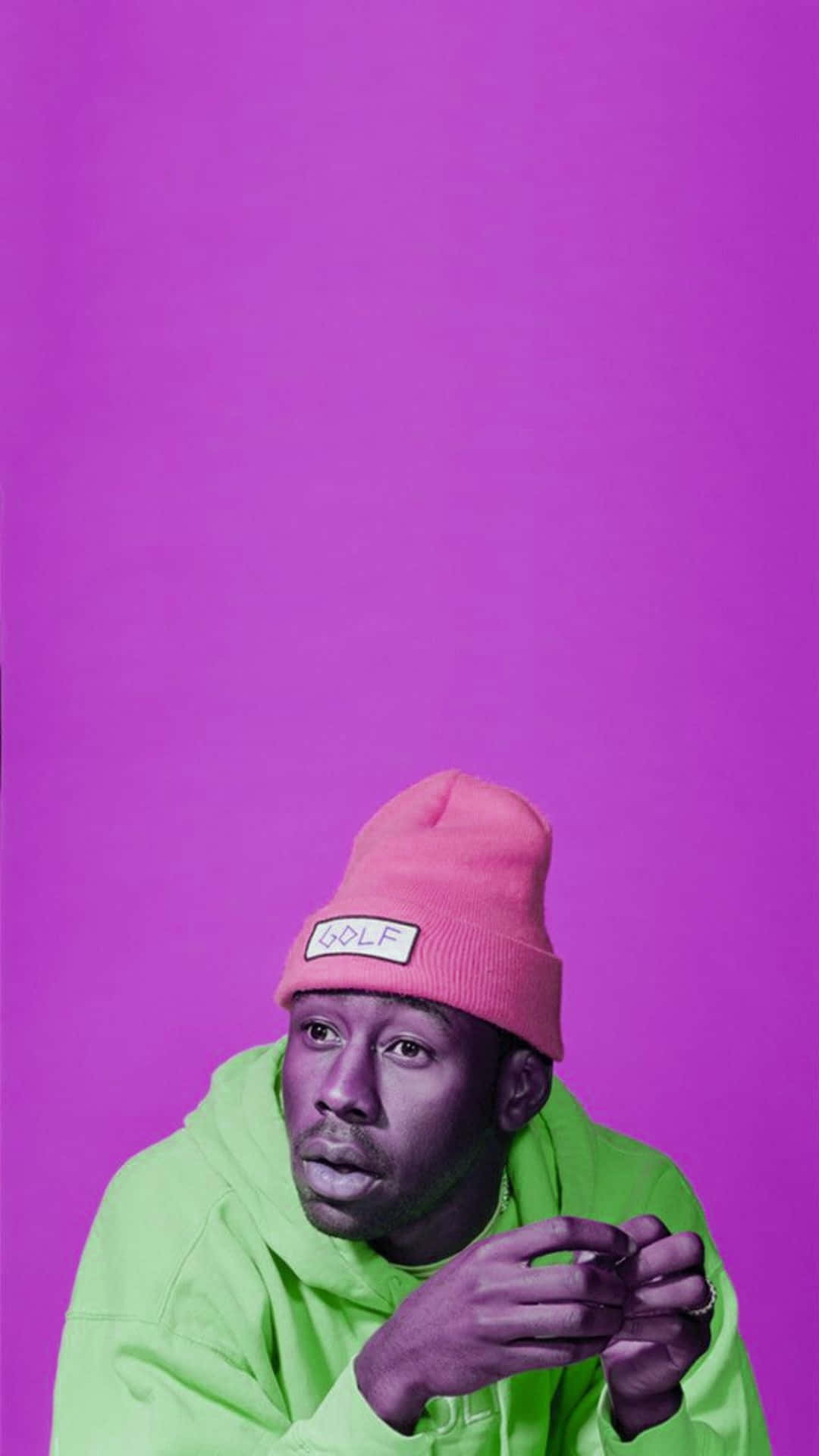 Tyler The Creator Aesthetic 桌布 壁紙 — 一個戴著粉紅色毛帽和螢光綠色連帽衫的男人，在純紫色背景前擺出沉思的表情。