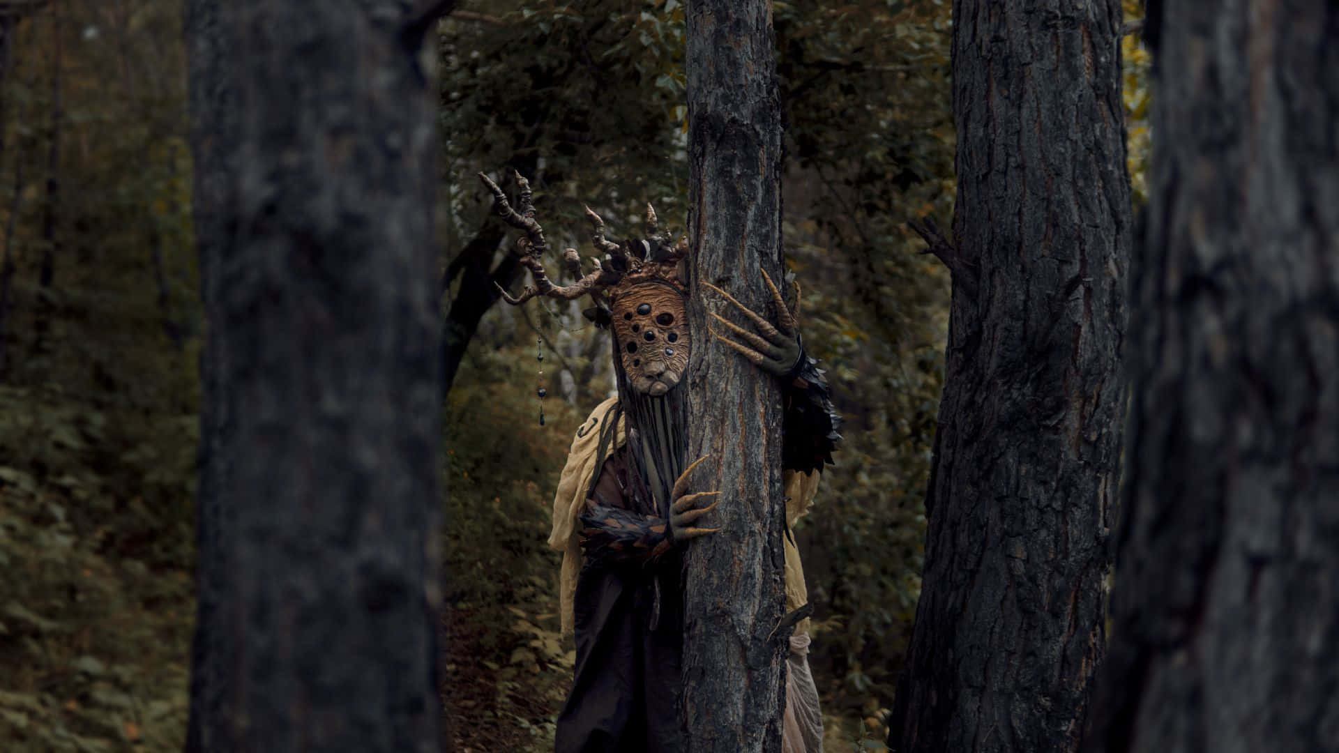 Horror Costumes 桌布 壁紙 — 一個戴著粗糙面具和鹿角的森林生物，在黑暗的林地中從樹幹後面窺視。