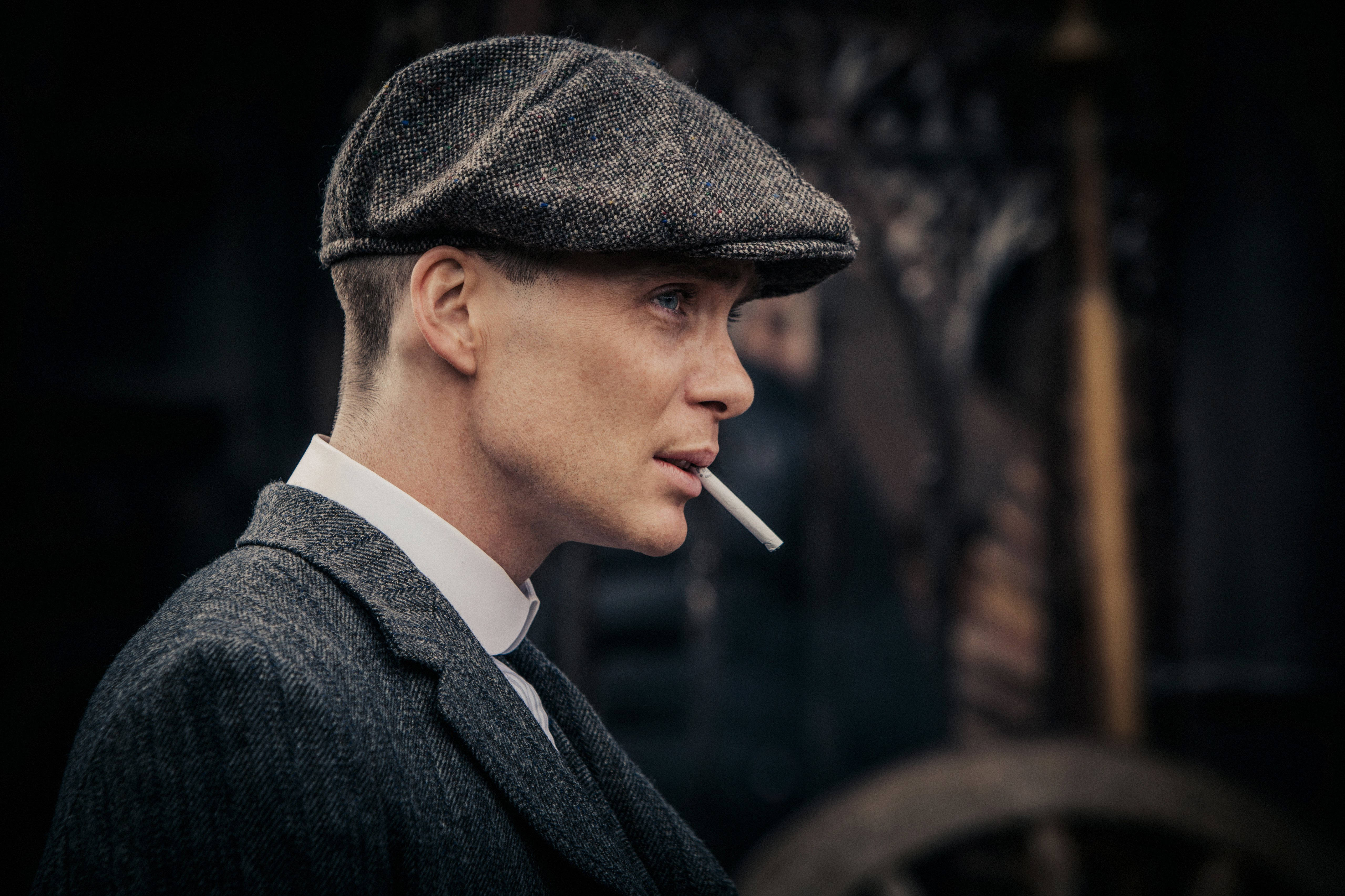 Thomas Shelby 桌布 壁紙 — 一個戴著粗花呢平頂帽、身穿西裝的男人在昏暗的復古環境中抽煙。