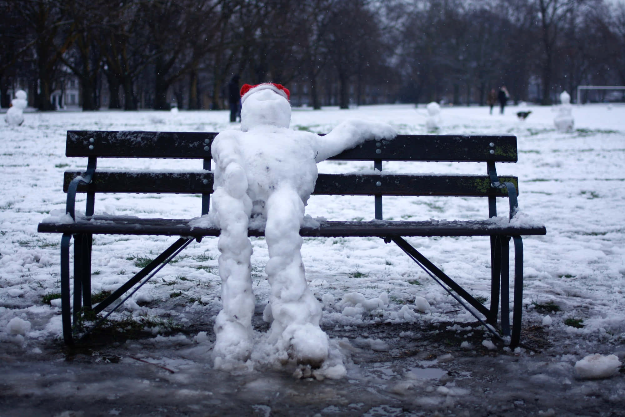 冬天 桌布 壁紙 — 一個戴著紅帽子的雪人坐在雪地公園的長椅上，周圍有光禿禿的樹木和飄落的雪花。