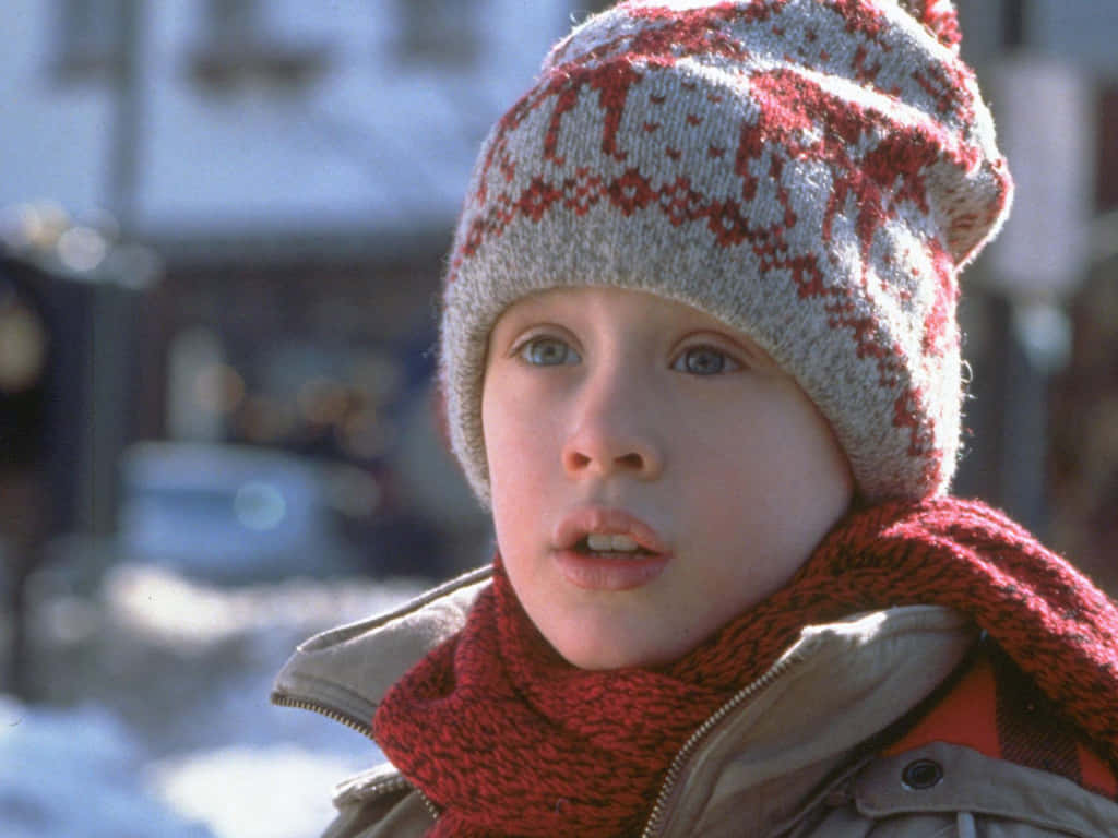 Home Alone 桌布 壁紙 — 一個戴著紅灰色針織帽和圍巾的小男孩，在白雪皚皚的城市背景下睜大眼睛凝視著。