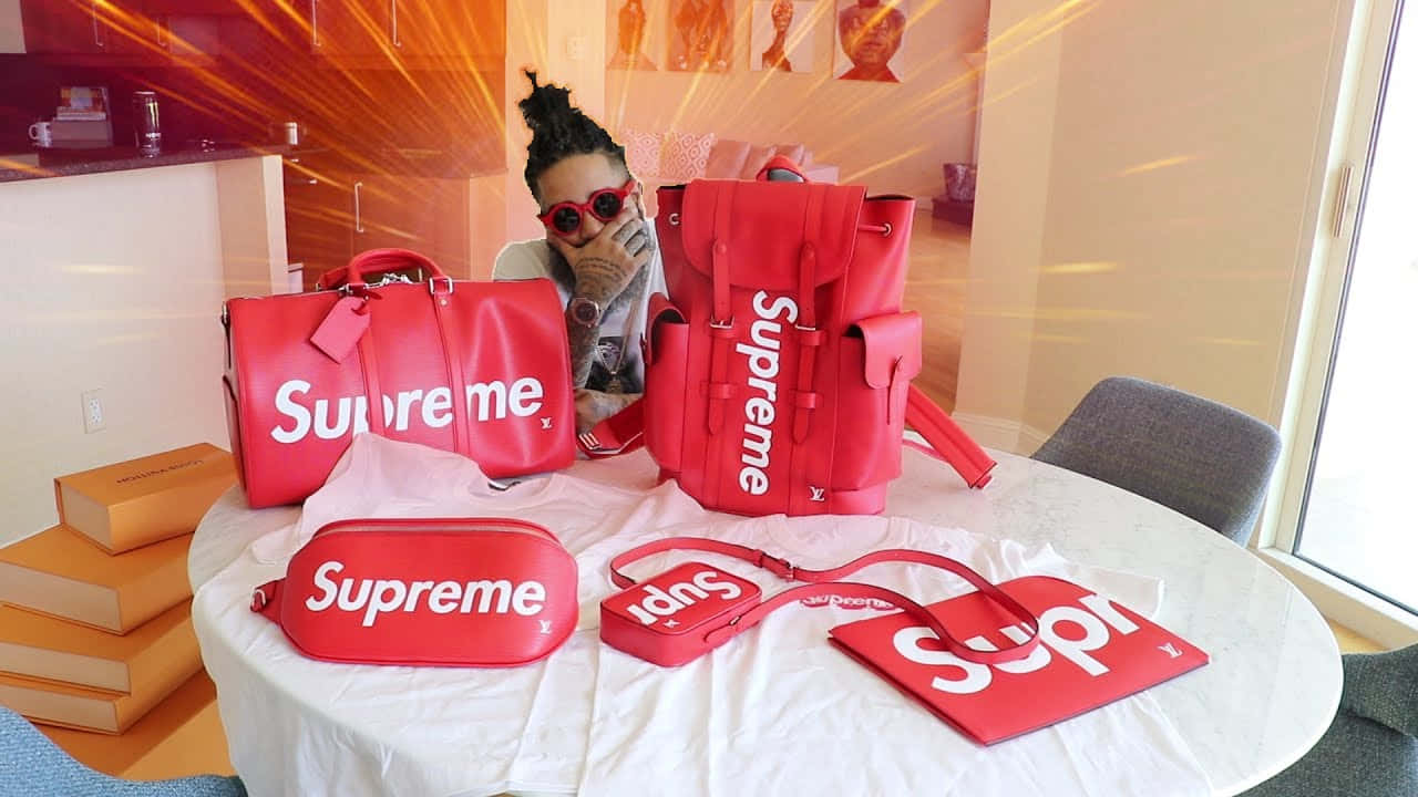 supreme 桌布 壁紙 — 一個戴著紅色太陽眼鏡的人在一個現代化的房間裡，在一張擺滿紅色 Supreme x Louis Vuitton 奢華手提袋和盒子的桌子後面微笑。