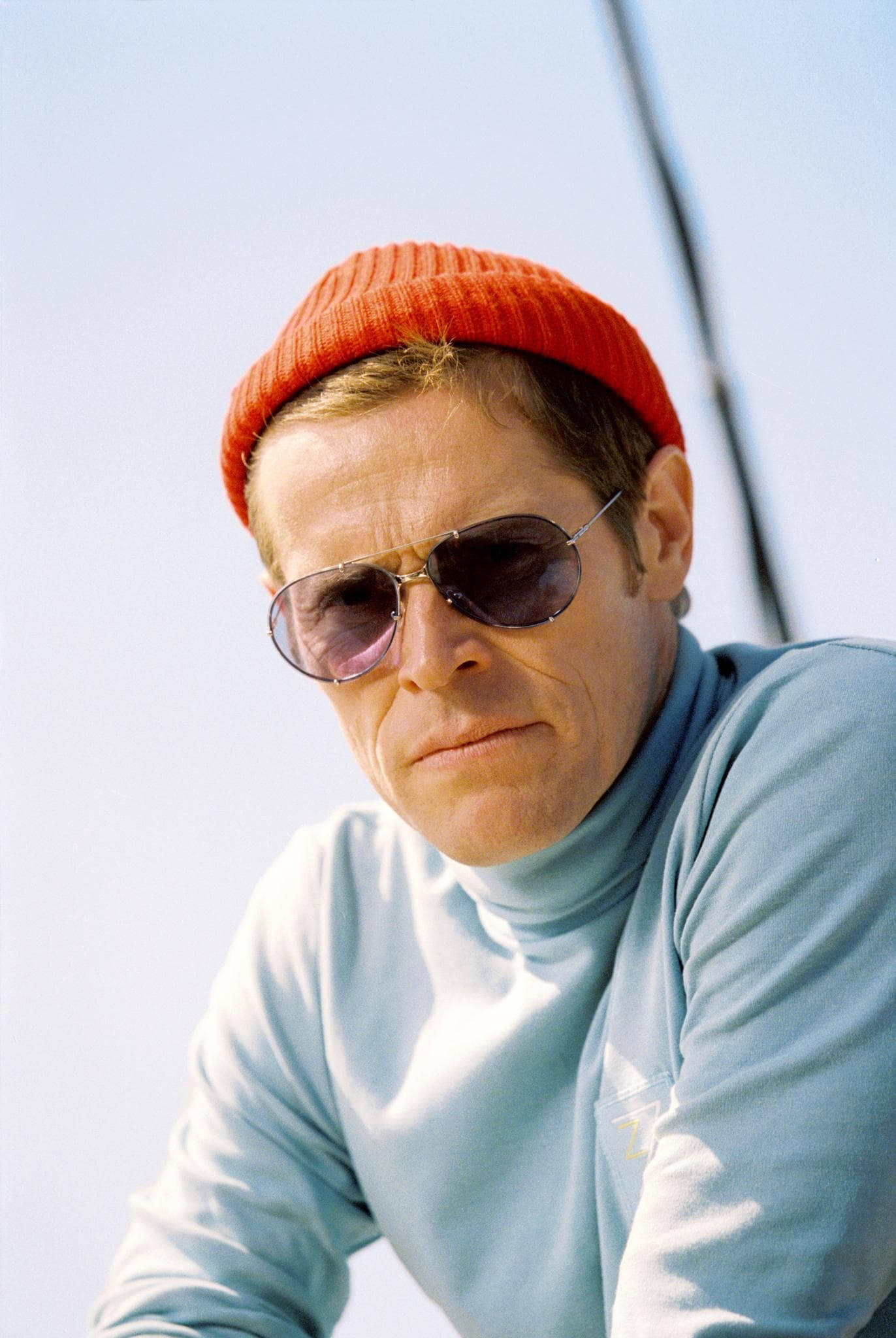 Willem Dafoe 桌布 壁紙 — 一個戴著紅色毛帽和太陽眼鏡的男人，在明亮的天空下凝視著，身後可見帆船的索具。