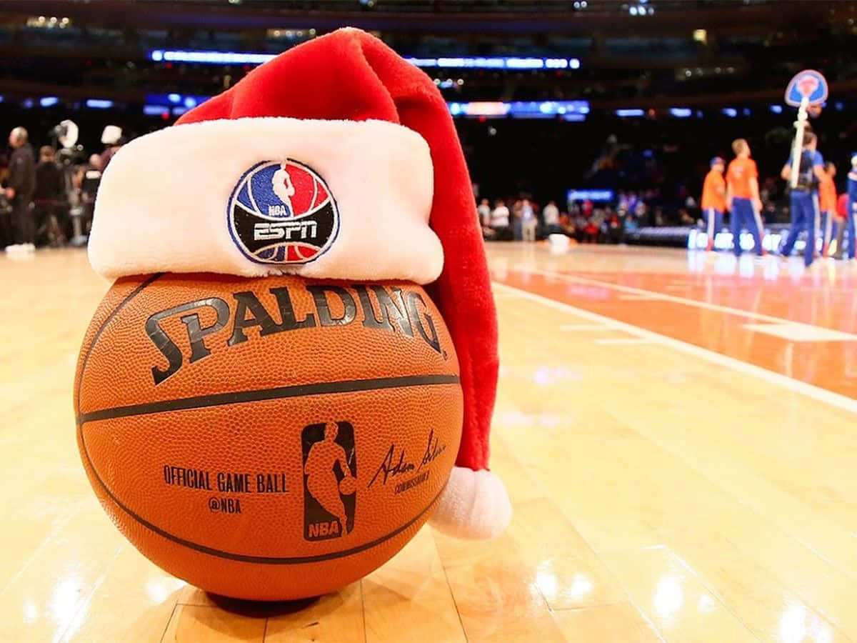 NBA聖誕節 桌布 壁紙 — 一個戴著紅色聖誕帽的Spalding NBA籃球，放在拋光的球場上，背景有球員和籃框。