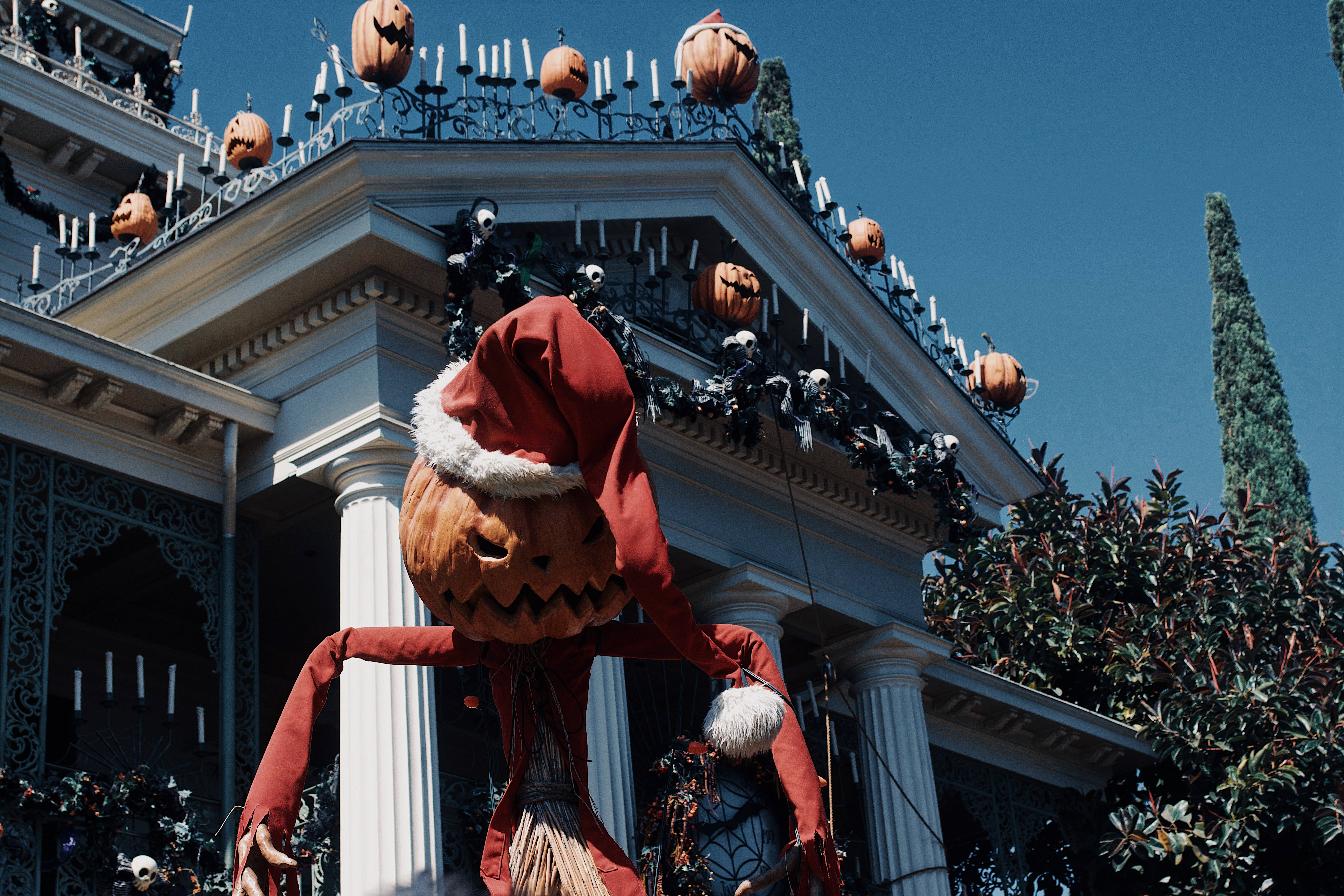 Disney Halloween 桌布 壁紙 — 一個戴著紅色聖誕帽的巨大南瓜燈站在一棟裝飾著南瓜和骷髏的大房子前，背景是晴朗的藍天。