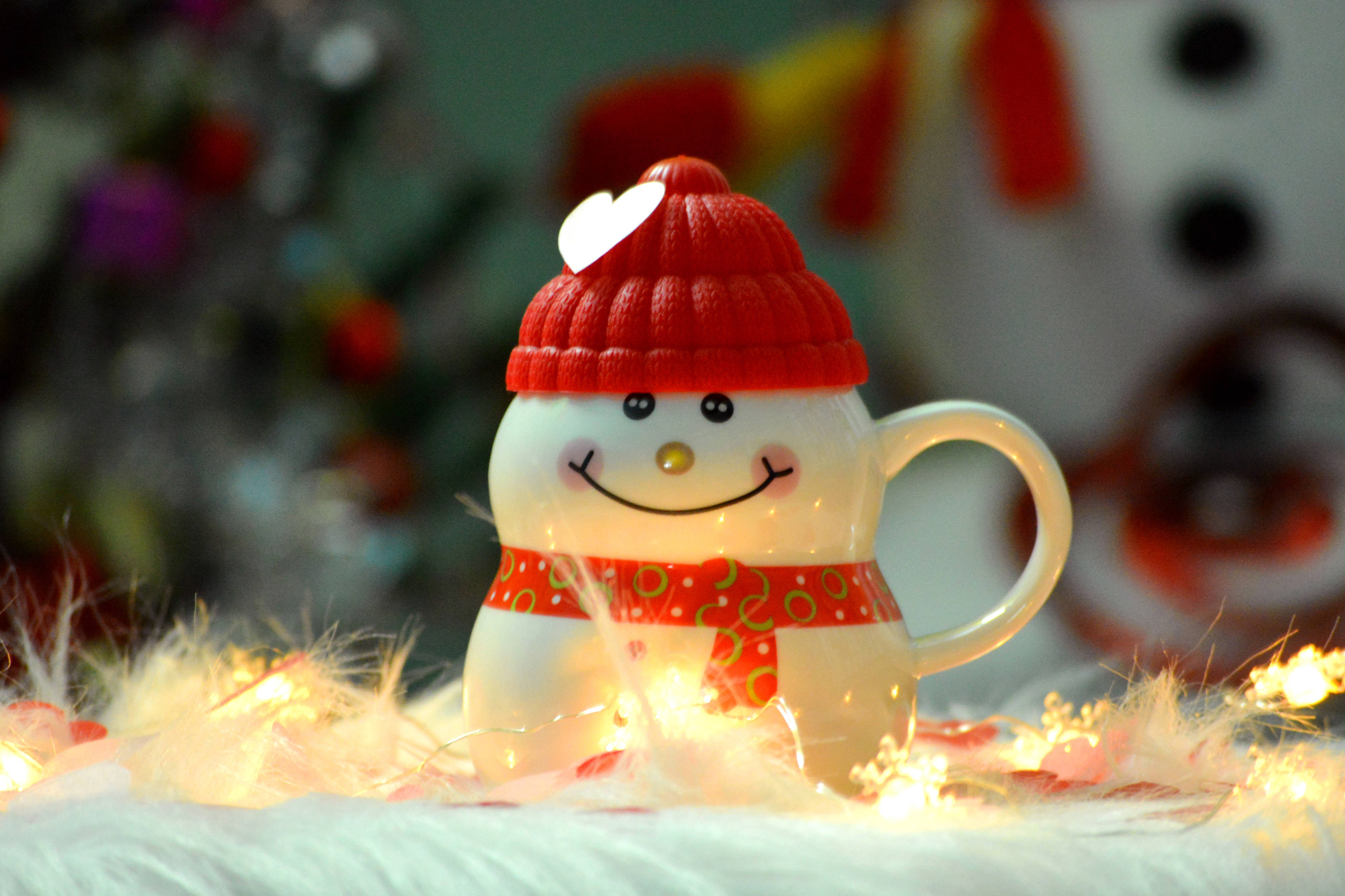 可愛聖誕快樂 桌布 壁紙 — 一個戴著紅色針織帽和圍巾的開心雪人馬克杯，坐在柔軟的白色毛絨上，周圍環繞著閃爍的仙女燈。
