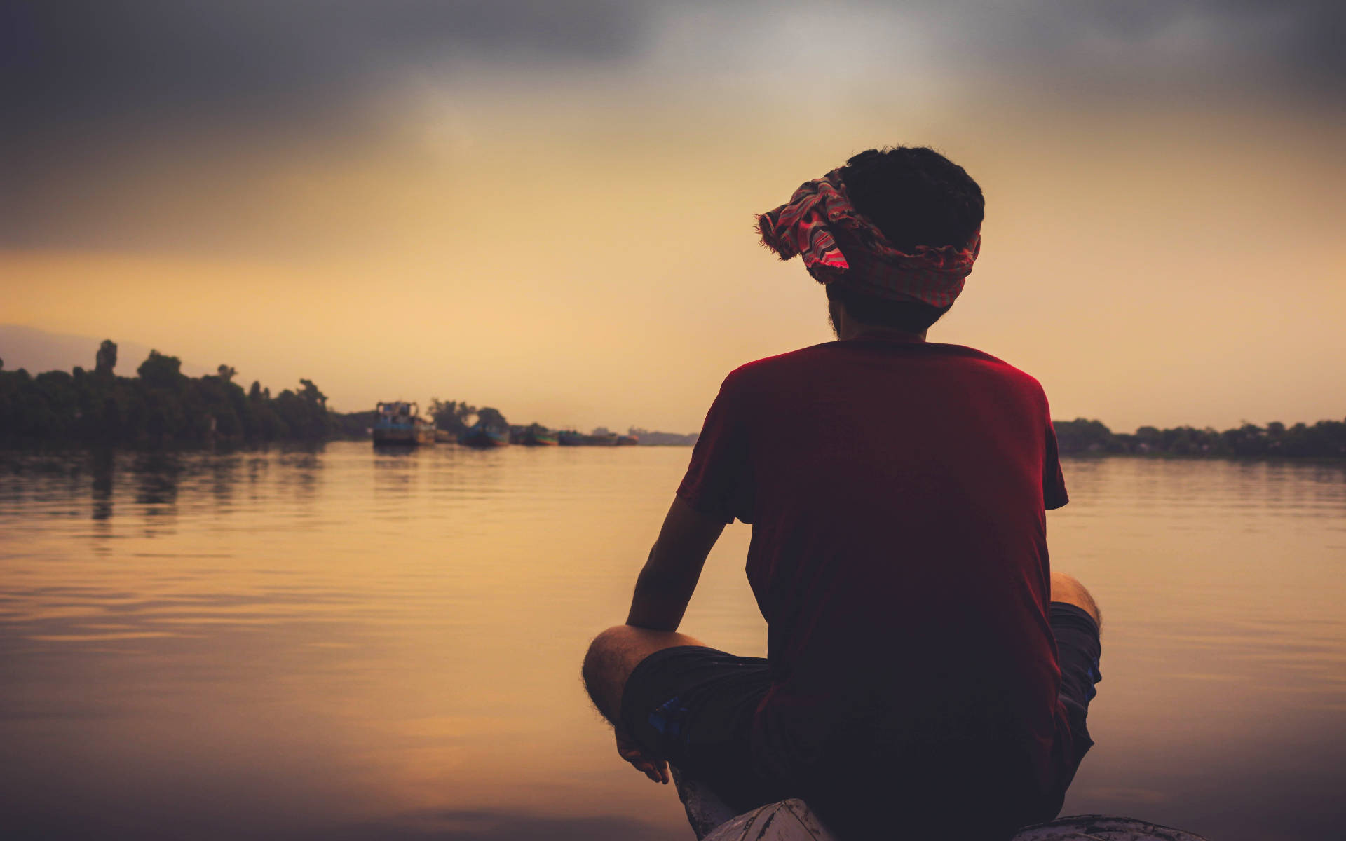 Alone 桌布 壁紙 — 一個戴著紅色頭巾的人坐在船上，在日落時分凝視著平靜的河流，河水映照著金橙色的天空。