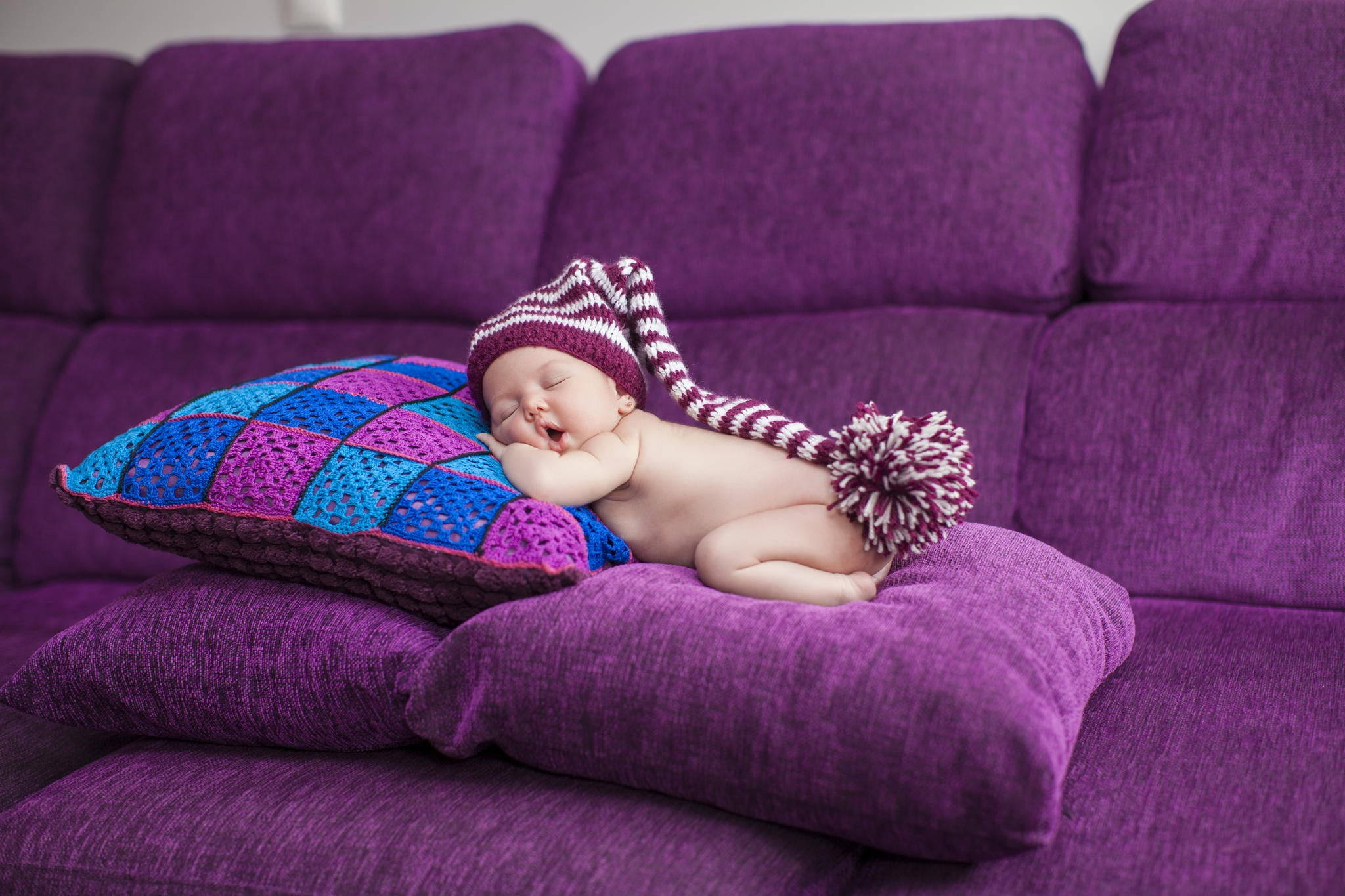 Baby Boy 桌布 壁紙 — 一個戴著紫白條紋針織帽（附有絨球）的熟睡新生嬰兒，安詳地躺在一個色彩繽紛的鉤織枕頭上，枕頭則放在一張柔軟的紫色沙發上。