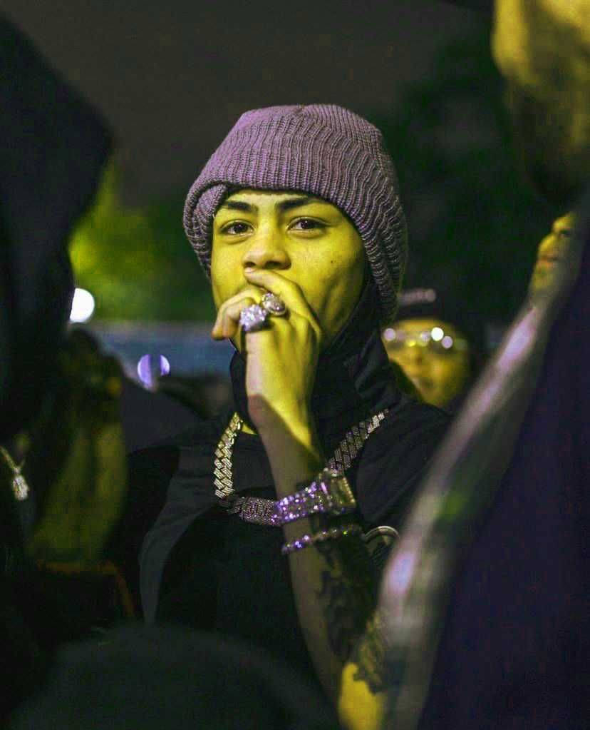 說唱歌手 桌布 壁紙 — 一個戴著紫色毛帽和黑色連帽衫的年輕男子在夜晚抽煙，周圍是模糊的人群和閃爍的燈光。