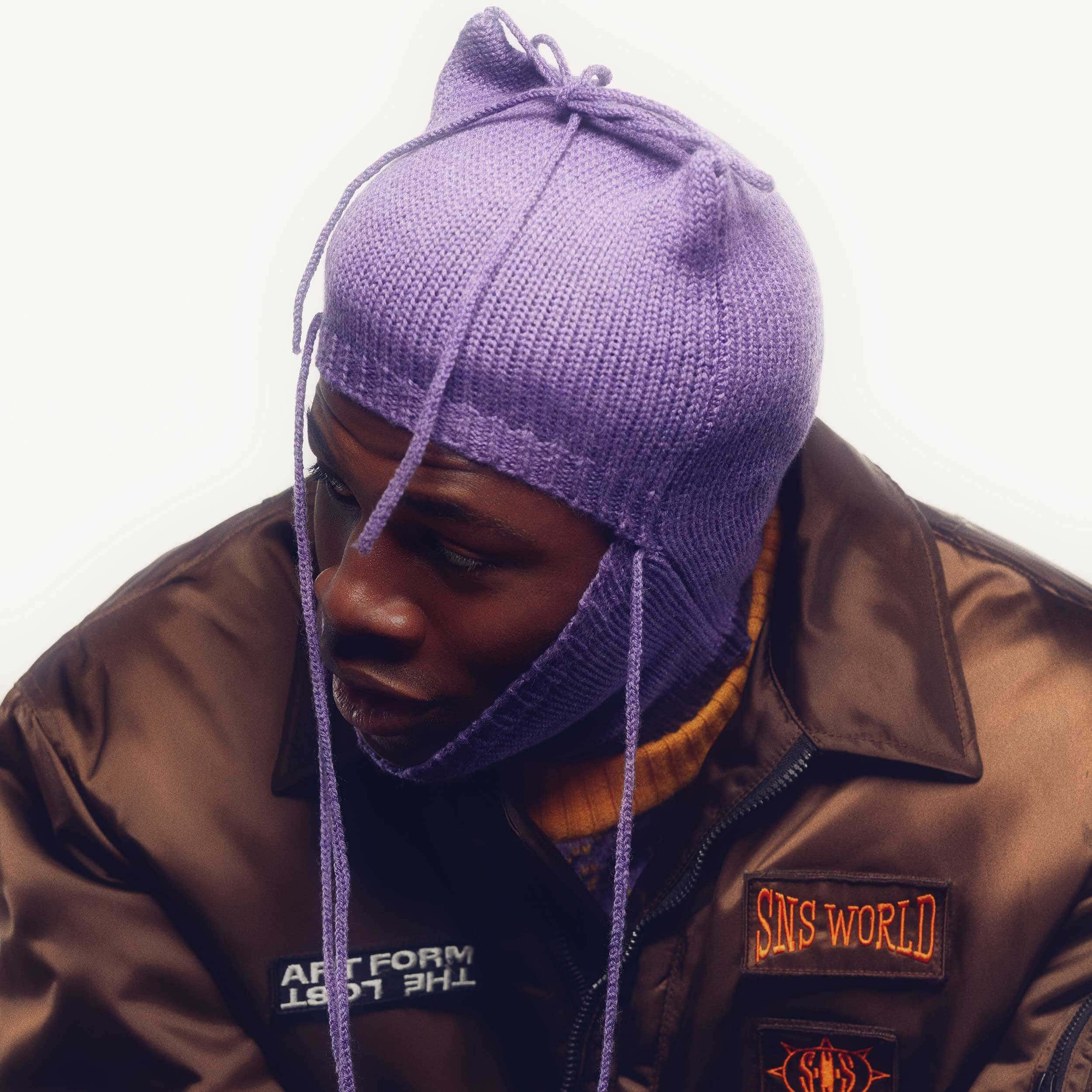 Purple Accessories 桌布 壁紙 — 一個戴著紫色針織毛帽和棕色夾克的男人，在純白色背景前低頭沉思。
