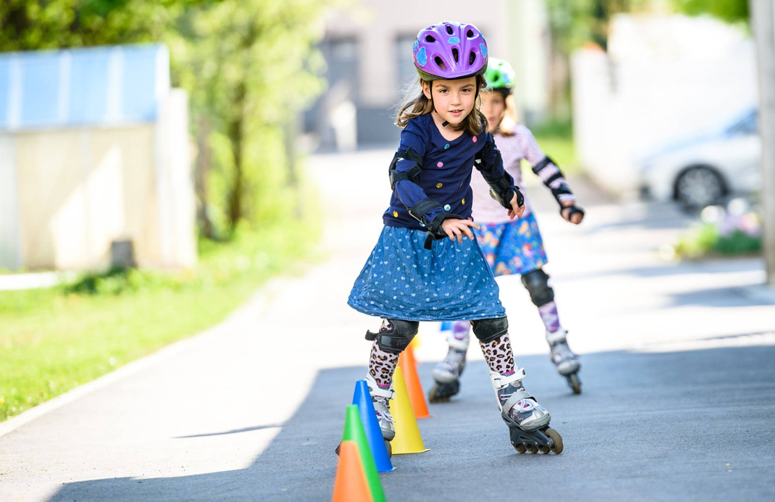 skating 桌布 壁紙 — 一個戴著紫色頭盔、穿著藍色連衣裙的小女孩在陽光明媚的郊區街道上自信地在彩色錐體之間溜冰。