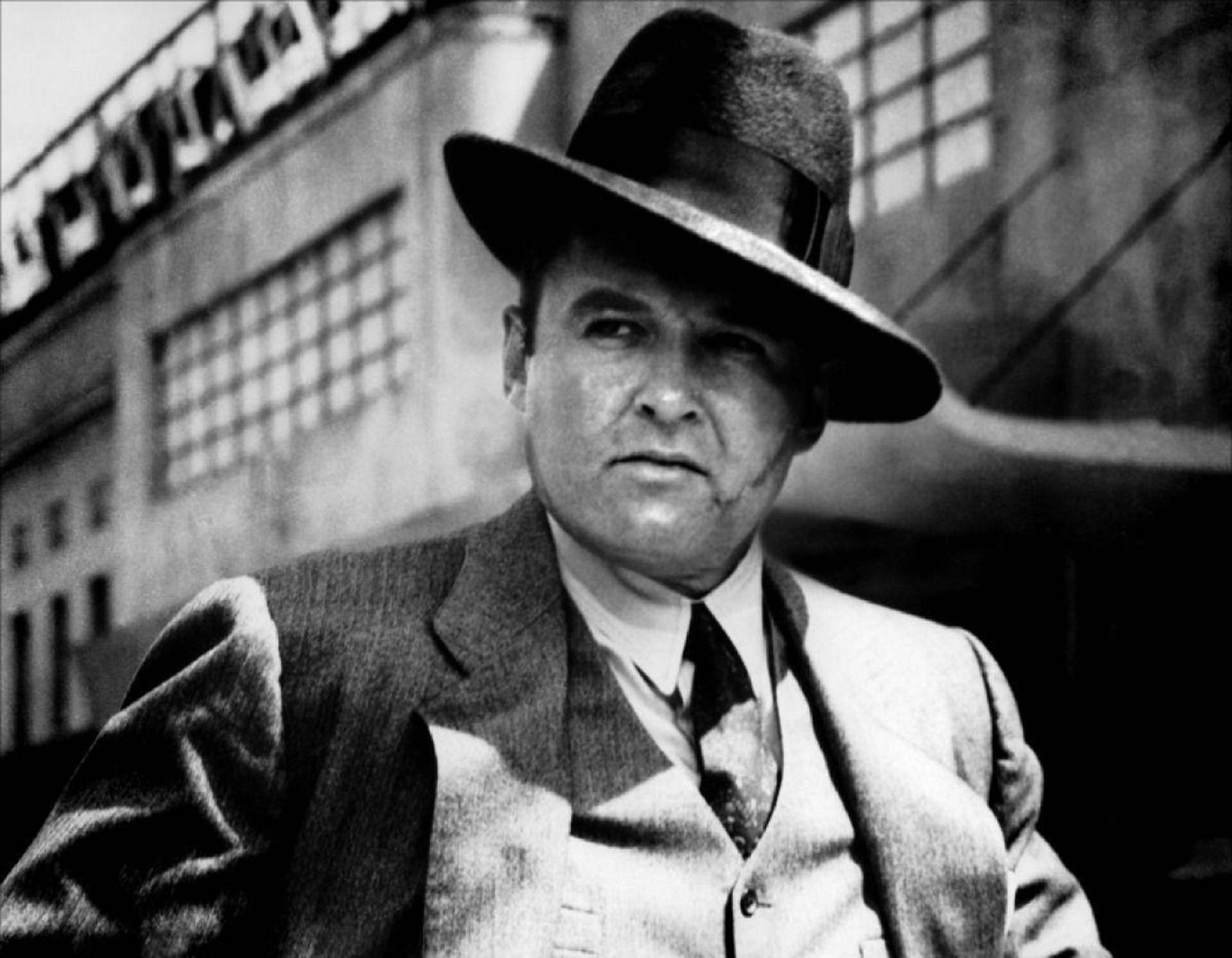Capone 桌布 壁紙 — 一個戴著紳士帽、穿著西裝的嚴肅男子，在經典黑白電影劇照中，對著粗糙的城市背景凝視。