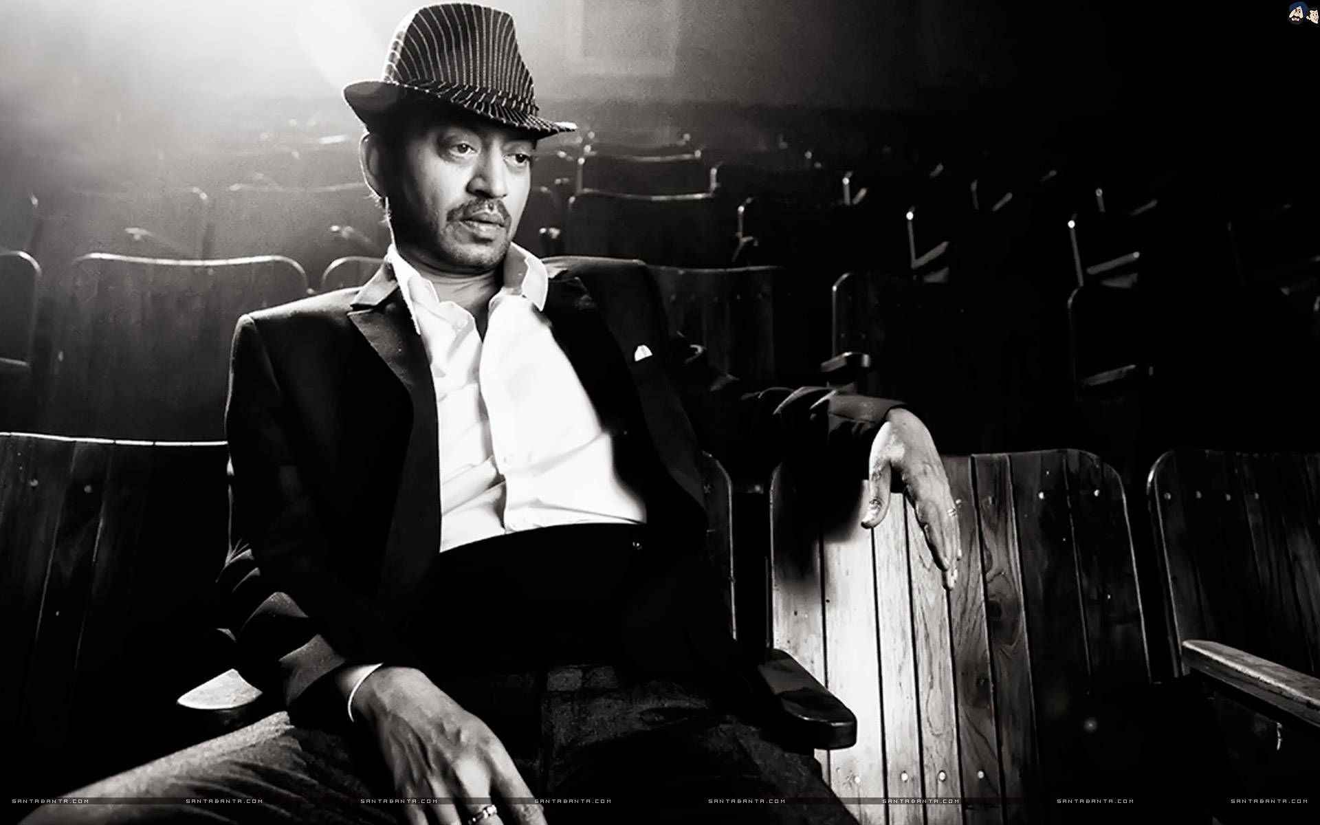 Irrfan Khan 桌布 壁紙 — 一個戴著紳士帽、穿著西裝的男人獨自坐在燈光昏暗的劇院中，背後有戲劇性的背光。
