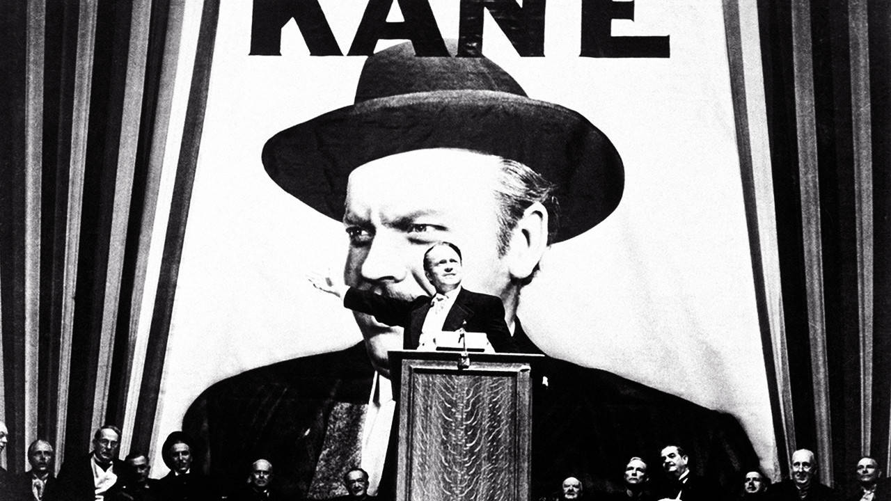 Citizen Kane 桌布 壁紙 — 一個戴著紳士帽的男人站在講台上，面前是一幅自己的巨大畫像，上方顯示著KANE的名字。