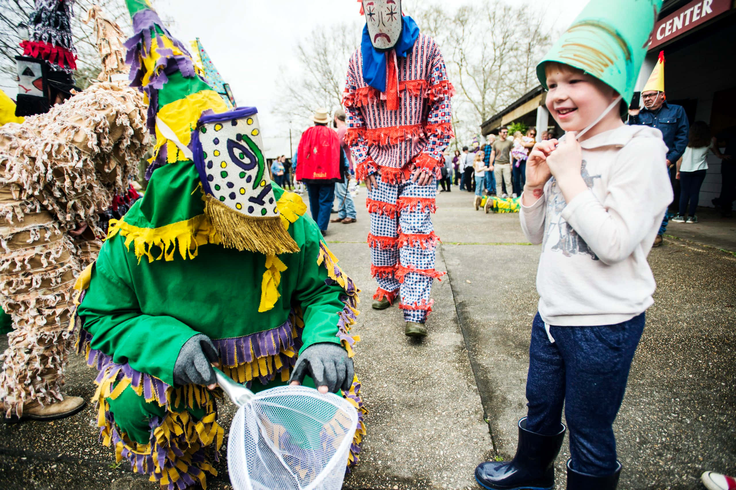 Mardi Gras 桌布 壁紙 — 一個戴著綠色帽子的快樂小孩微笑著，看著色彩繽紛、戴著面具的狂歡者穿著節日服裝在街上遊行。