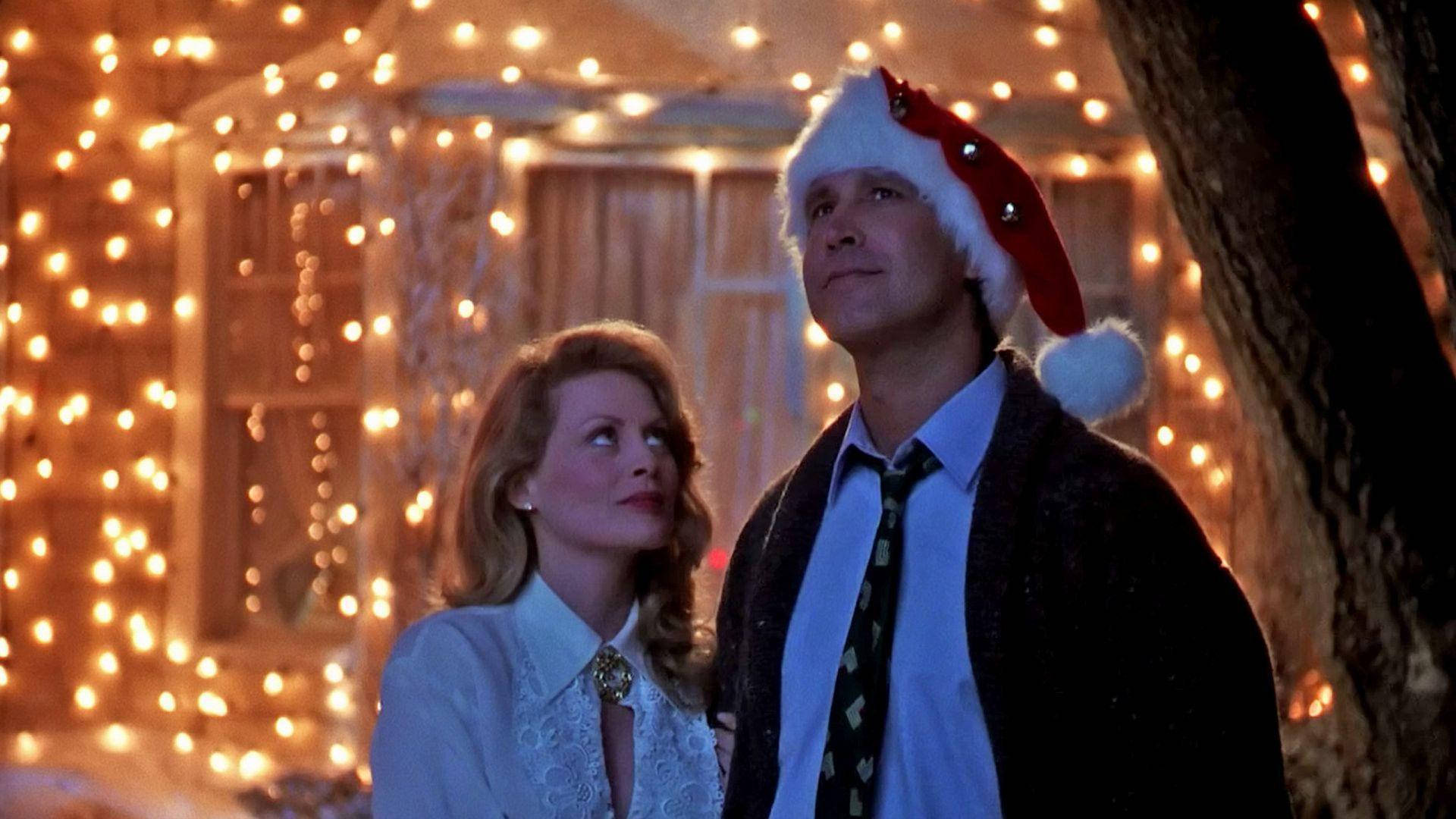 Christmas Vacation 桌布 壁紙 — 一個戴著聖誕帽的男人和一個女人在溫暖的燈串光芒中向上凝望，背景是白雪皚皚的冬日場景。