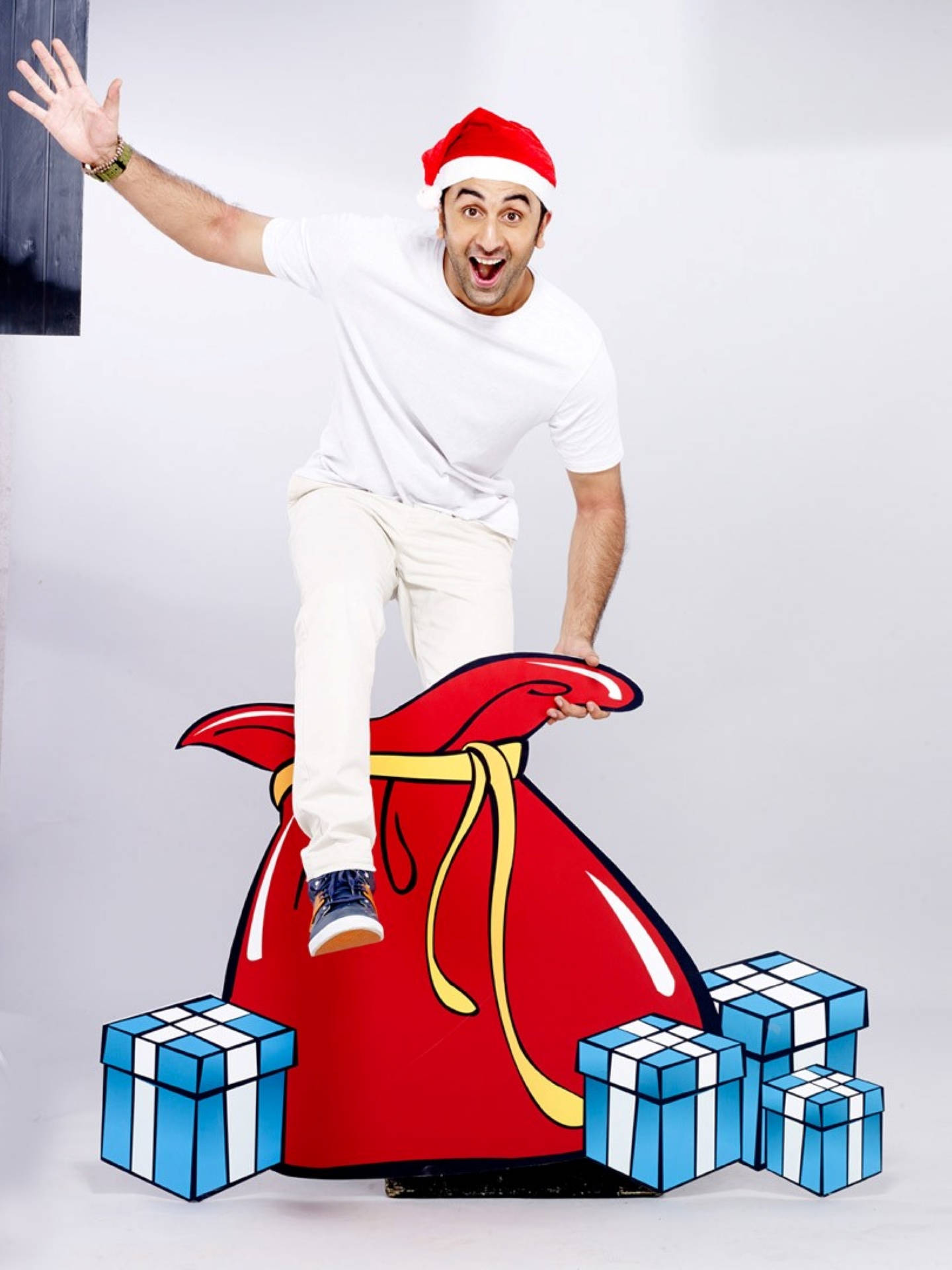 Ranbir Kapoor 桌布 壁紙 — 一個戴著聖誕帽的男人愉快地平衡在一個巨大的紅色禮物袋上，周圍環繞著藍色和白色的禮物。