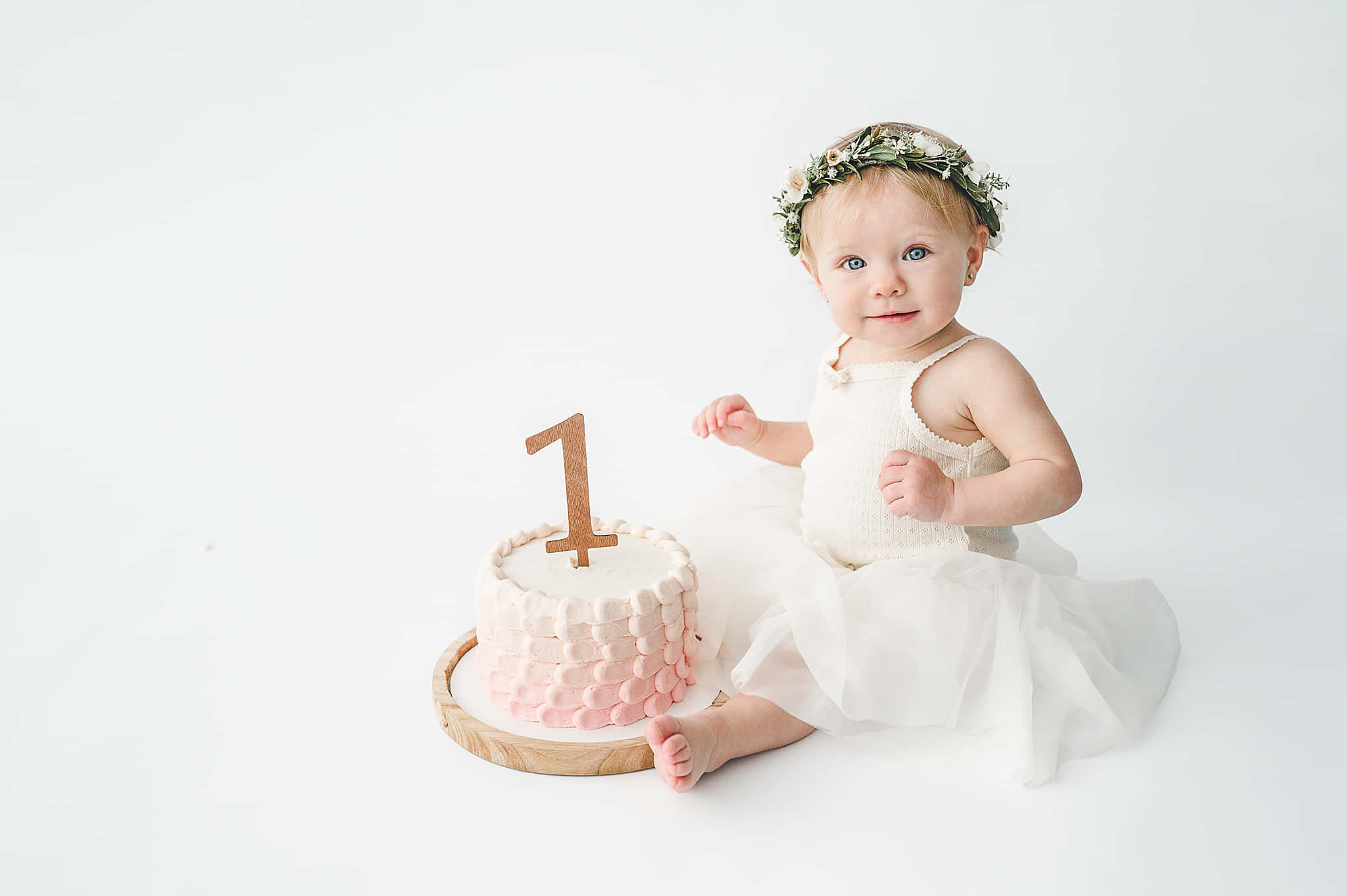 Birthday Cake 桌布 壁紙 — 一個戴著花冠、身穿白色連衣裙的女嬰坐在白色背景上，旁邊是一個粉紅色紋理蛋糕，上面有一個木製的數字一裝飾。
