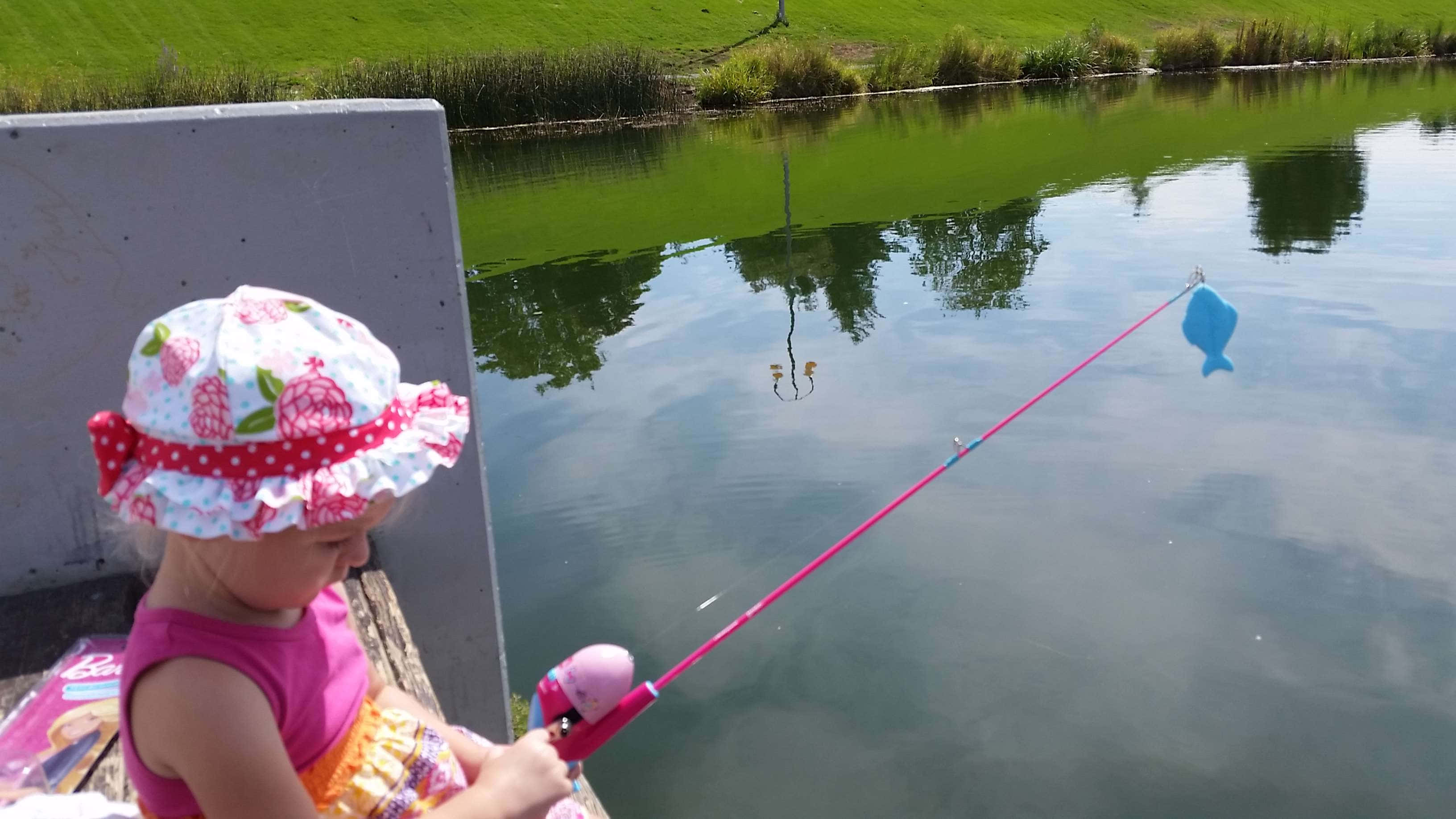 Fishing Rod 桌布 壁紙 — 一個戴著花卉帽的小孩在陽光明媚的日子裡，將粉紅色的釣竿拋入平靜的綠色池塘中。