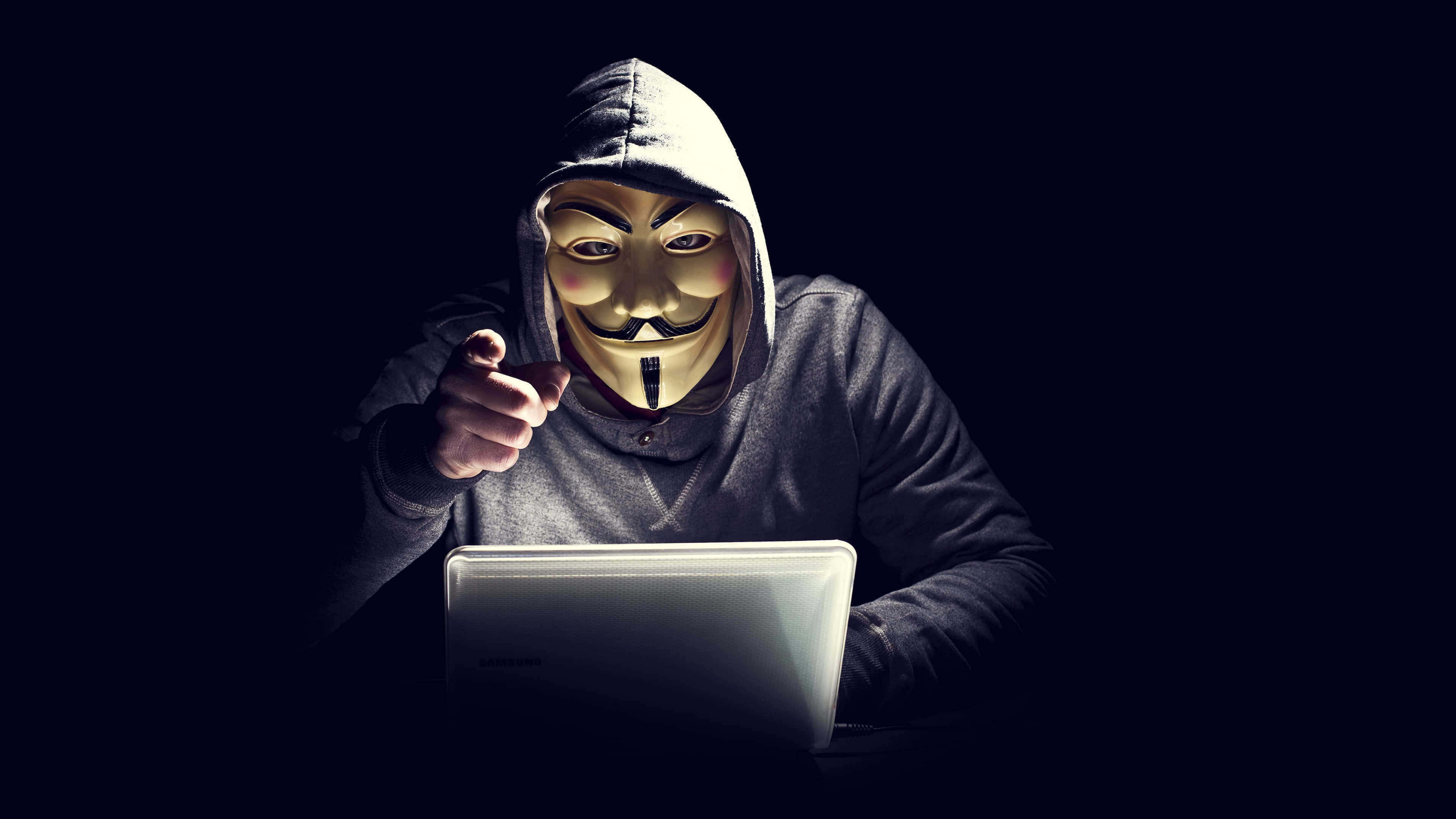 匿名 桌布 壁紙 — 一個戴著蓋伊·福克斯面具和連帽衫的人，在黑暗的房間裡，被螢幕的光芒照亮，指責地指著一台筆記型電腦。