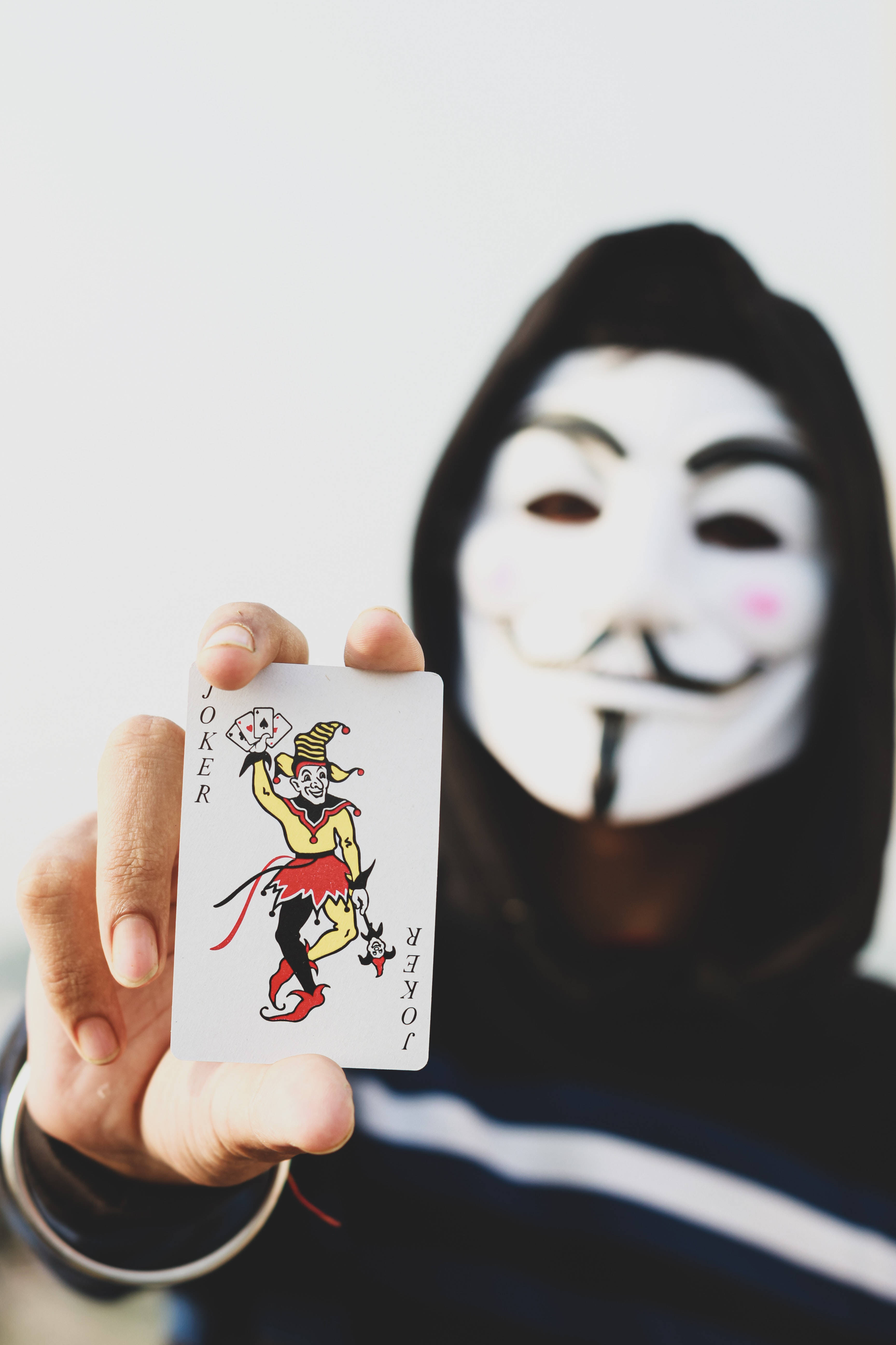 駭客面具 桌布 壁紙 — 一個戴著蓋伊·福克斯面具的人，在純白色背景前舉起一張小丑撲克牌。
