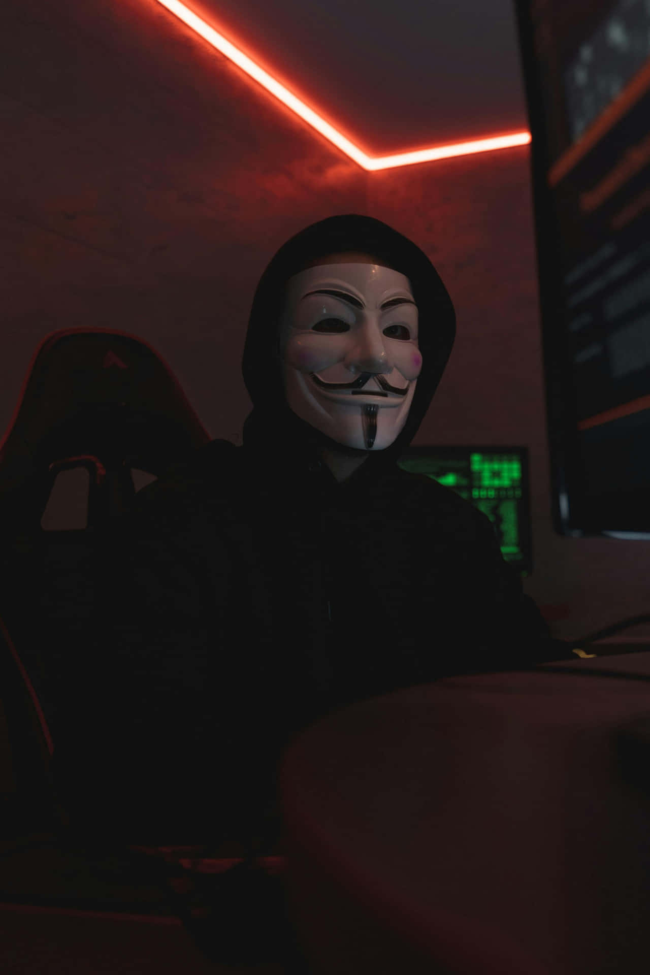  壁紙 — 一個戴著蓋伊·福克斯面具的人坐在一個昏暗的房間裡，房間被天花板的紅色霓虹燈條和發光的電腦螢幕淹沒。