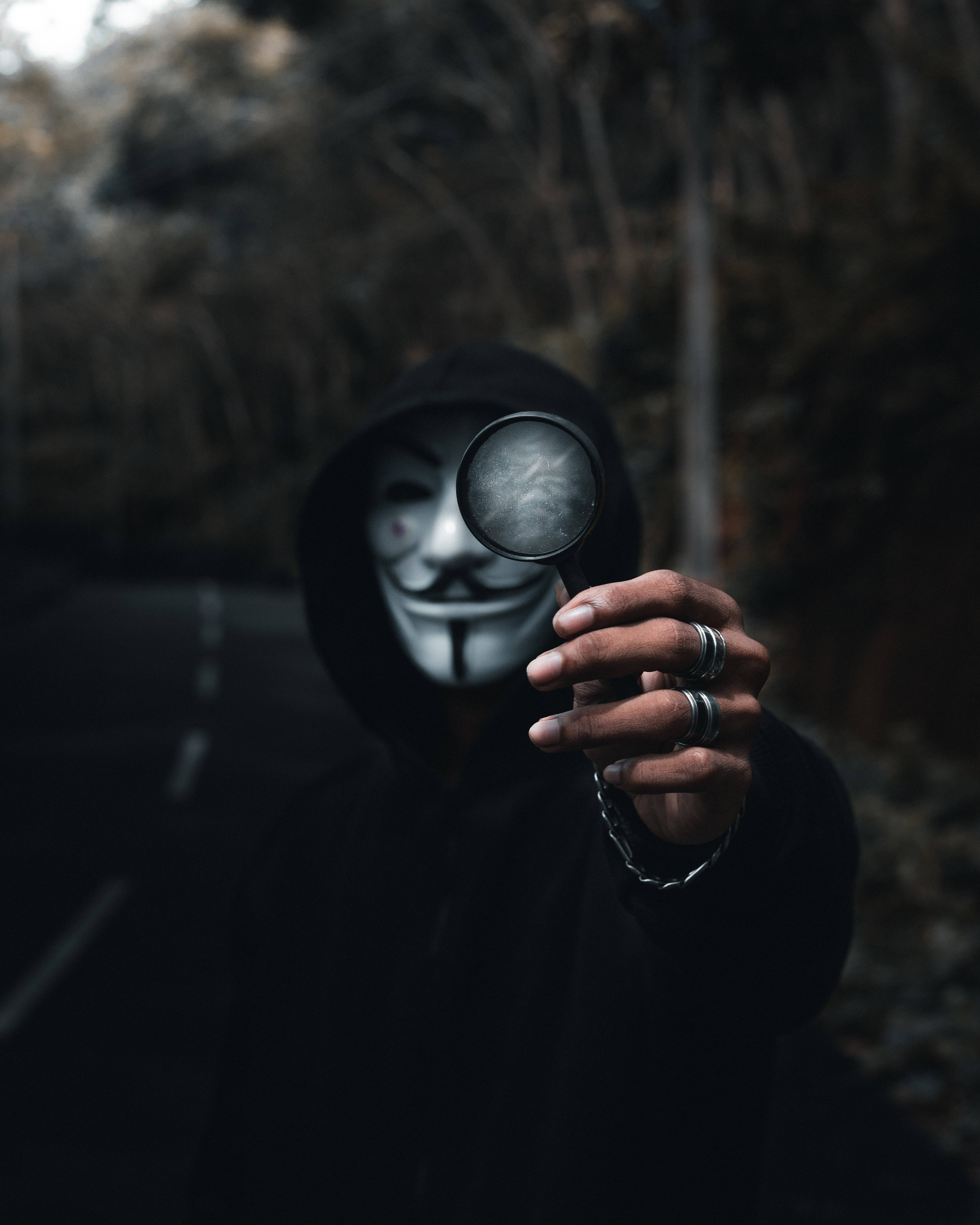 匿名 桌布 壁紙 — 一個戴著蓋伊·福克斯面具的兜帽人物在昏暗的森林道路上向觀看者舉起放大鏡。