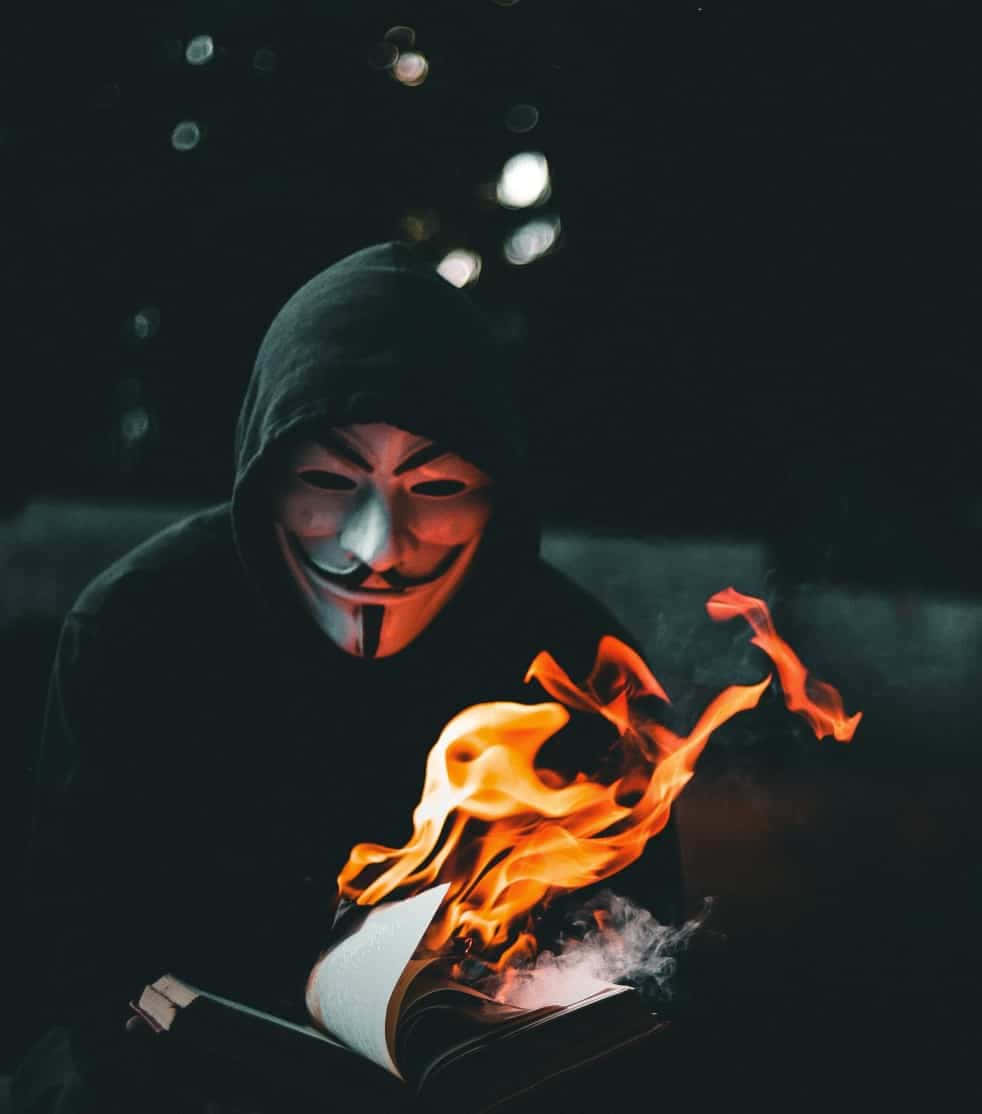 高清 桌布 壁紙 — 一個戴著蓋伊·福克斯面具的兜帽人物，在黑暗背景和散景燈光下，看著火焰吞噬一本打開的書。