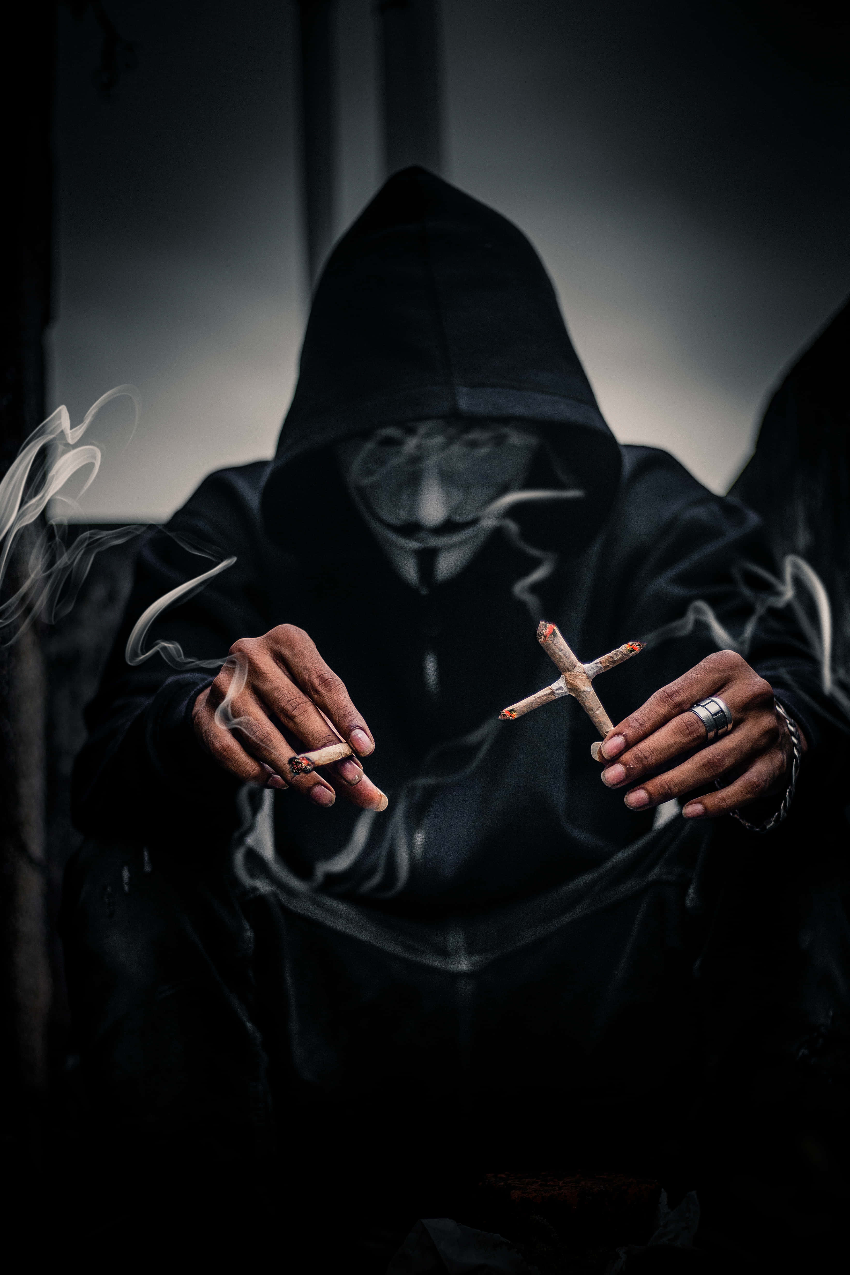 Mask 桌布 壁紙 — 一個戴著蓋伊·福克斯面具的連帽人物在昏暗燈光下抽著十字形大麻煙，同時手持一根香煙。