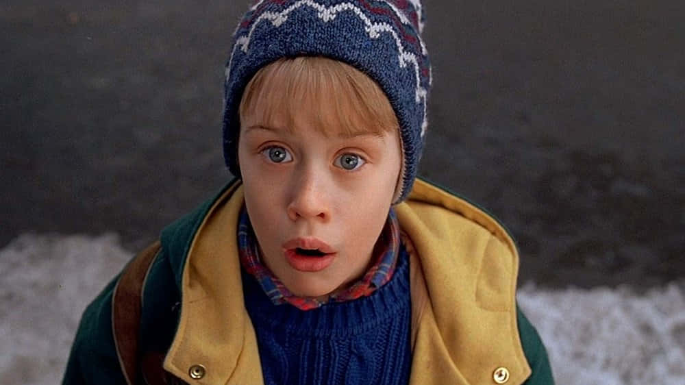 Home Alone 桌布 壁紙 — 一個戴著藍色毛帽、穿著黃色背心的小男孩，在雪地背景下瞪大眼睛，一臉震驚。