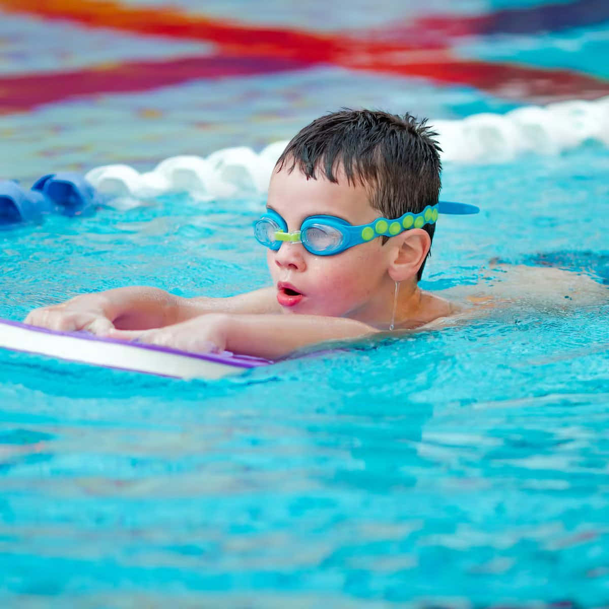 Swimming 桌布 壁紙 — 一個戴著藍色泳鏡和紫色浮板的男孩，在明亮的藍色泳池中專注地游泳。