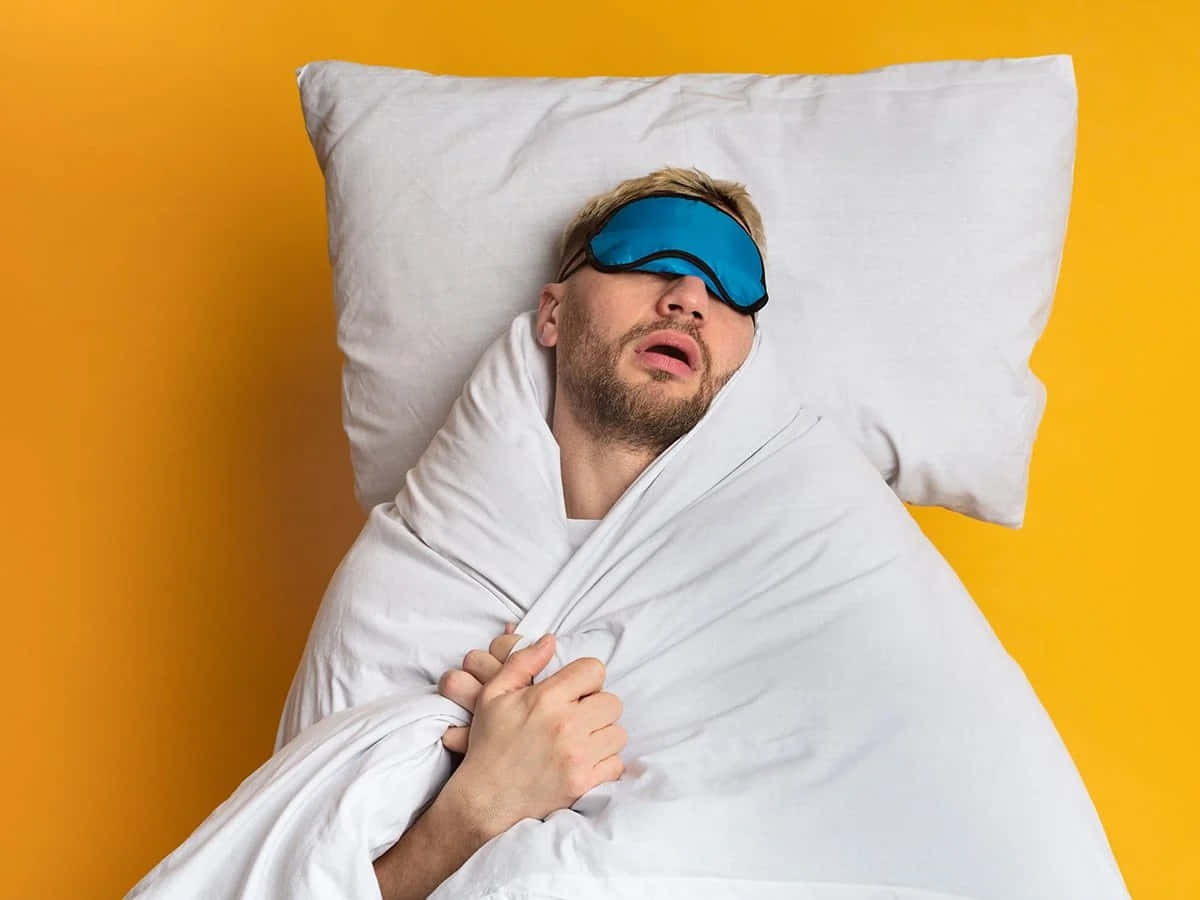 搞笑 桌布 壁紙 — 一個戴著藍色睡眠眼罩的男人，蓋著白色被子，枕著白色枕頭，在鮮黃色的牆壁前安然入睡。