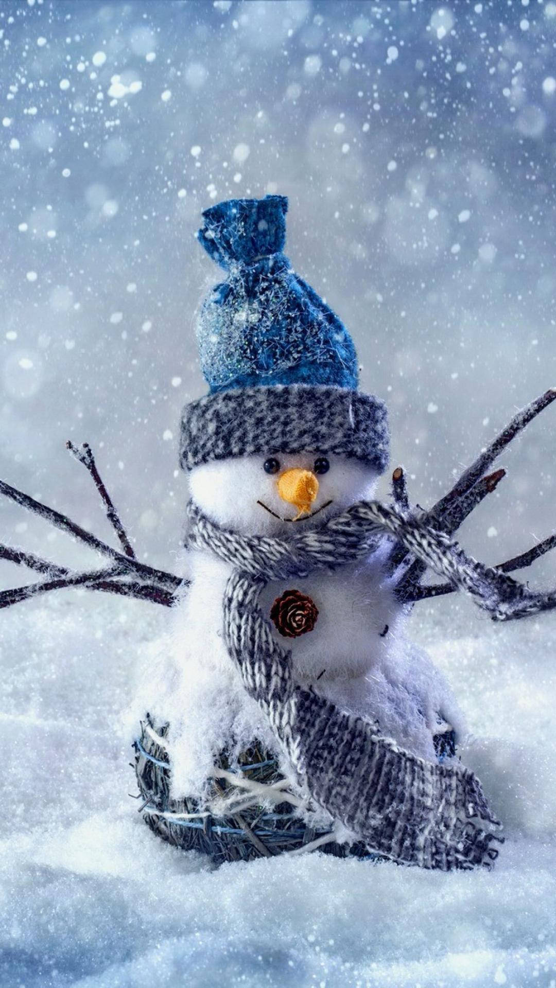 Cute Winter iPhone 桌布 壁紙 — 一個戴著藍色針織帽和圍巾的快樂雪人，用樹枝手臂站在飄落的雪中。