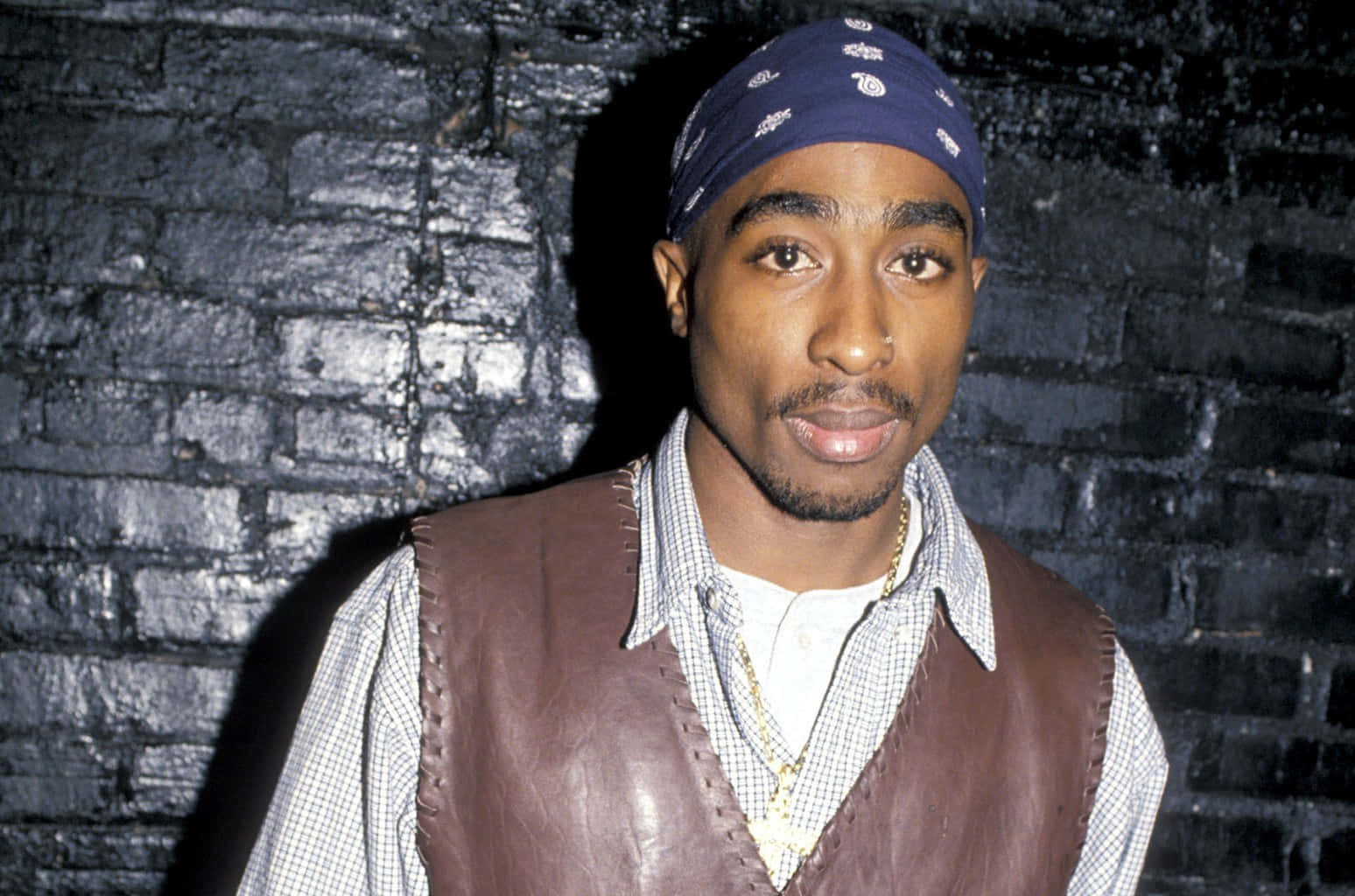 tupac 桌布 壁紙 — 一個戴著藍色頭巾和棕色皮背心的男人，在戲劇性的燈光下站在深色磚牆前。