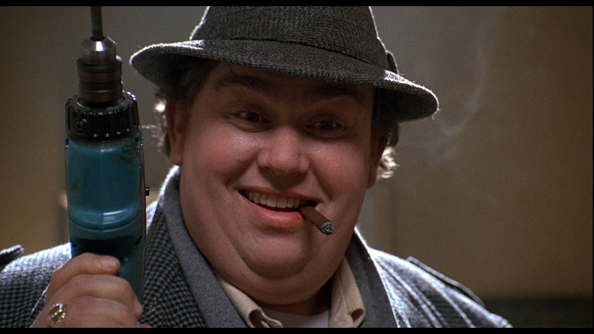 John Candy 桌布 壁紙 — 一個戴著費多拉帽、穿著千鳥格大衣的男人咧嘴笑著，一手拿著藍色電鑽，一手抽著雪茄。