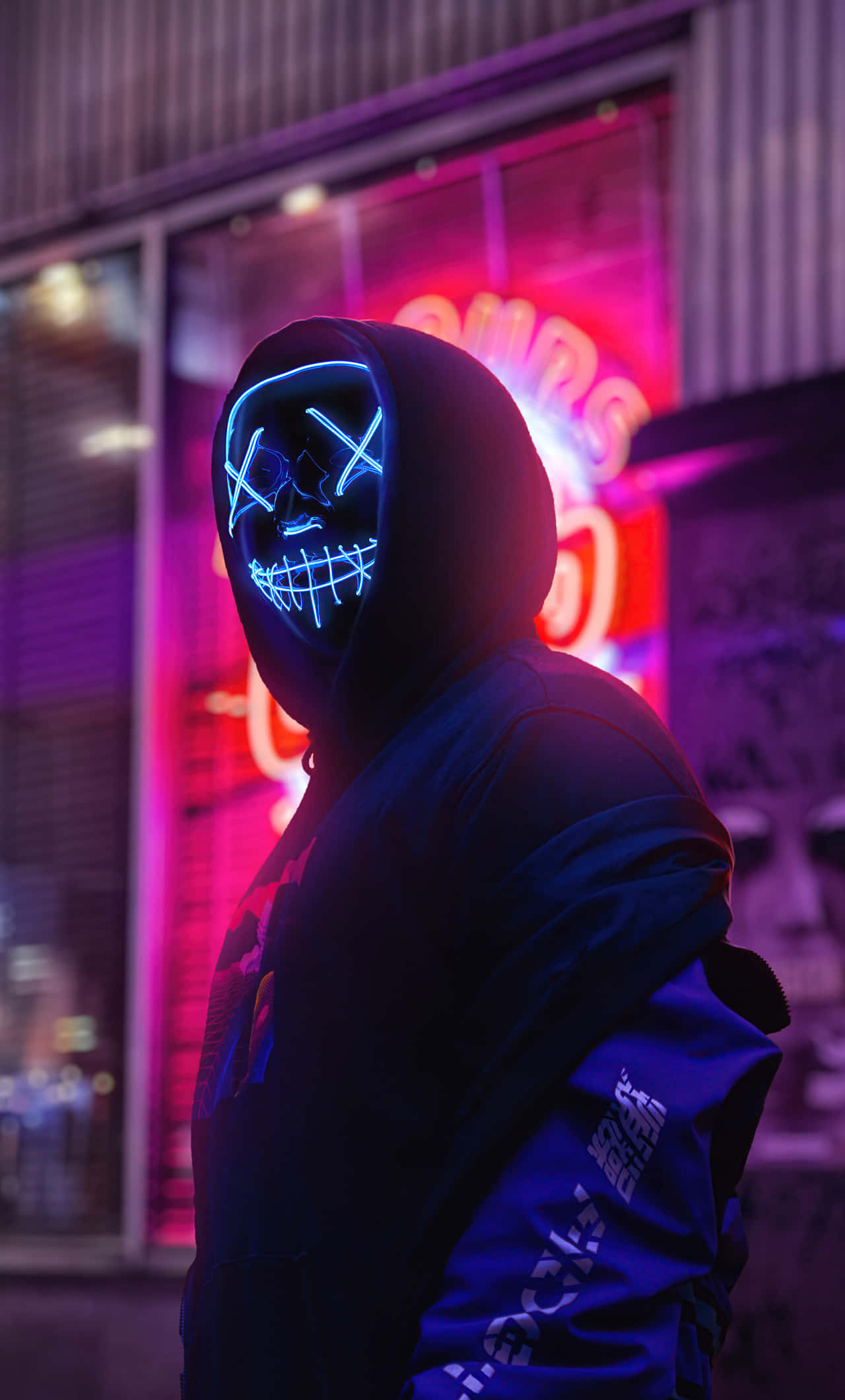 Mask Boy 桌布 壁紙 — 一個戴著連帽的人物戴著發光的藍色骷髏面具，背景是充滿活力的紫色和粉紅色霓虹燈城市景觀。