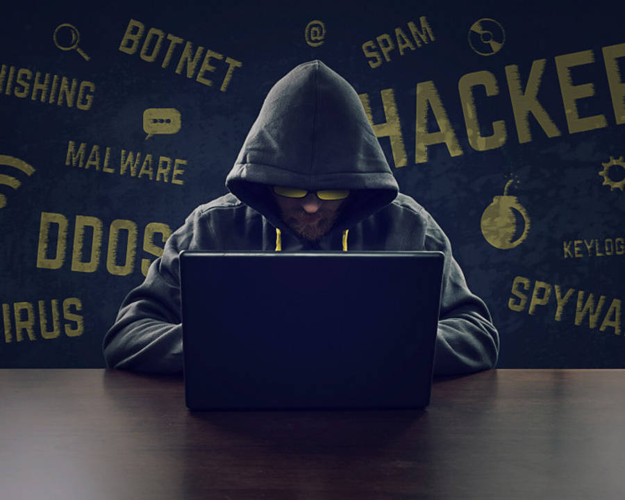 hacker 3d 桌布 壁紙 — 一個戴著連帽衫、戴著發光眼鏡的黑客在筆記型電腦上打字，周圍的暗牆上寫滿了惡意軟體和殭屍網絡等網絡犯罪詞語。