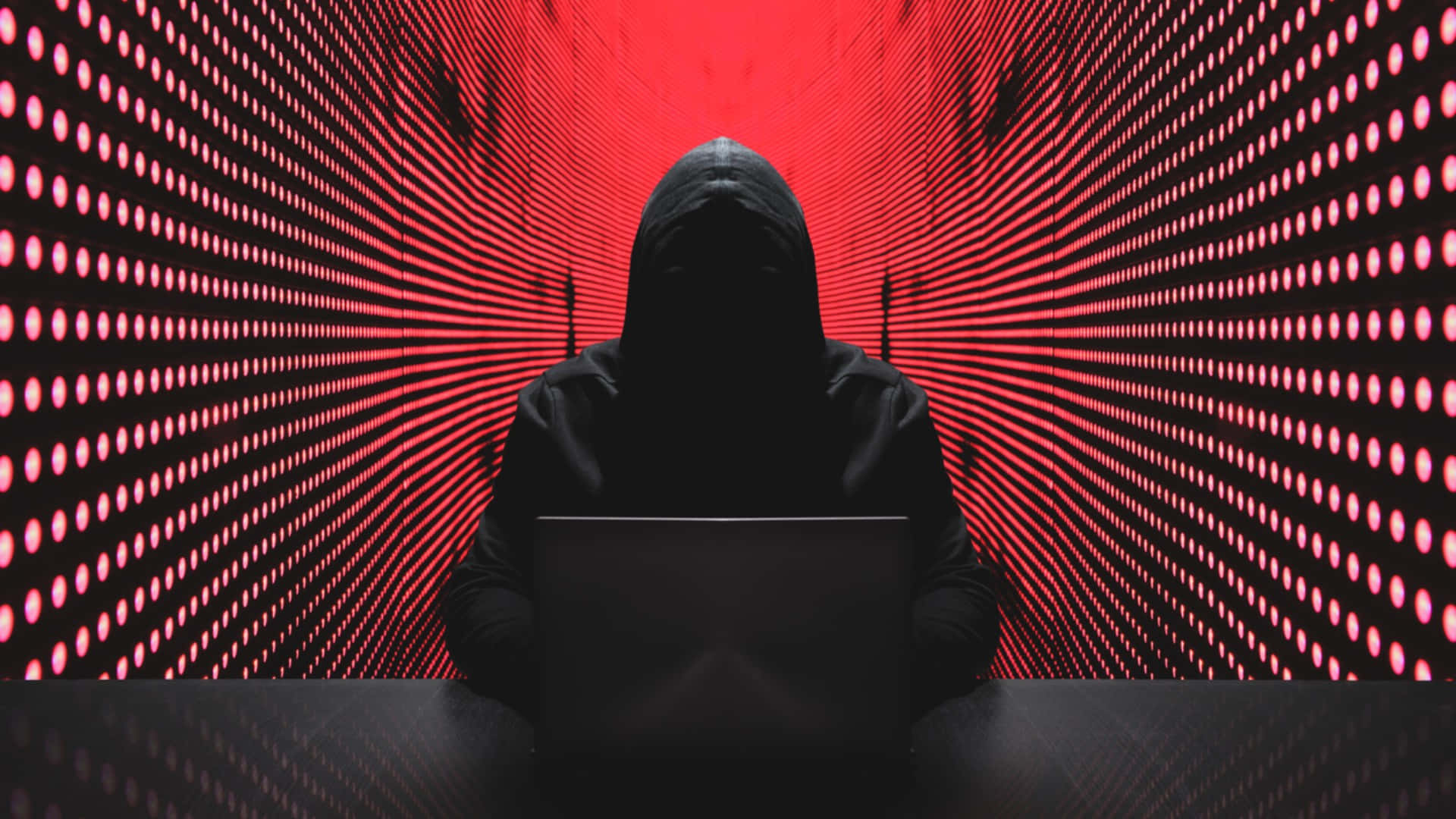 Hacking 桌布 壁紙 — 一個戴著連帽衫的人物坐在陰影中，面前是一台筆記型電腦，背景是脈動的紅色數位隧道，佈滿發光的點。