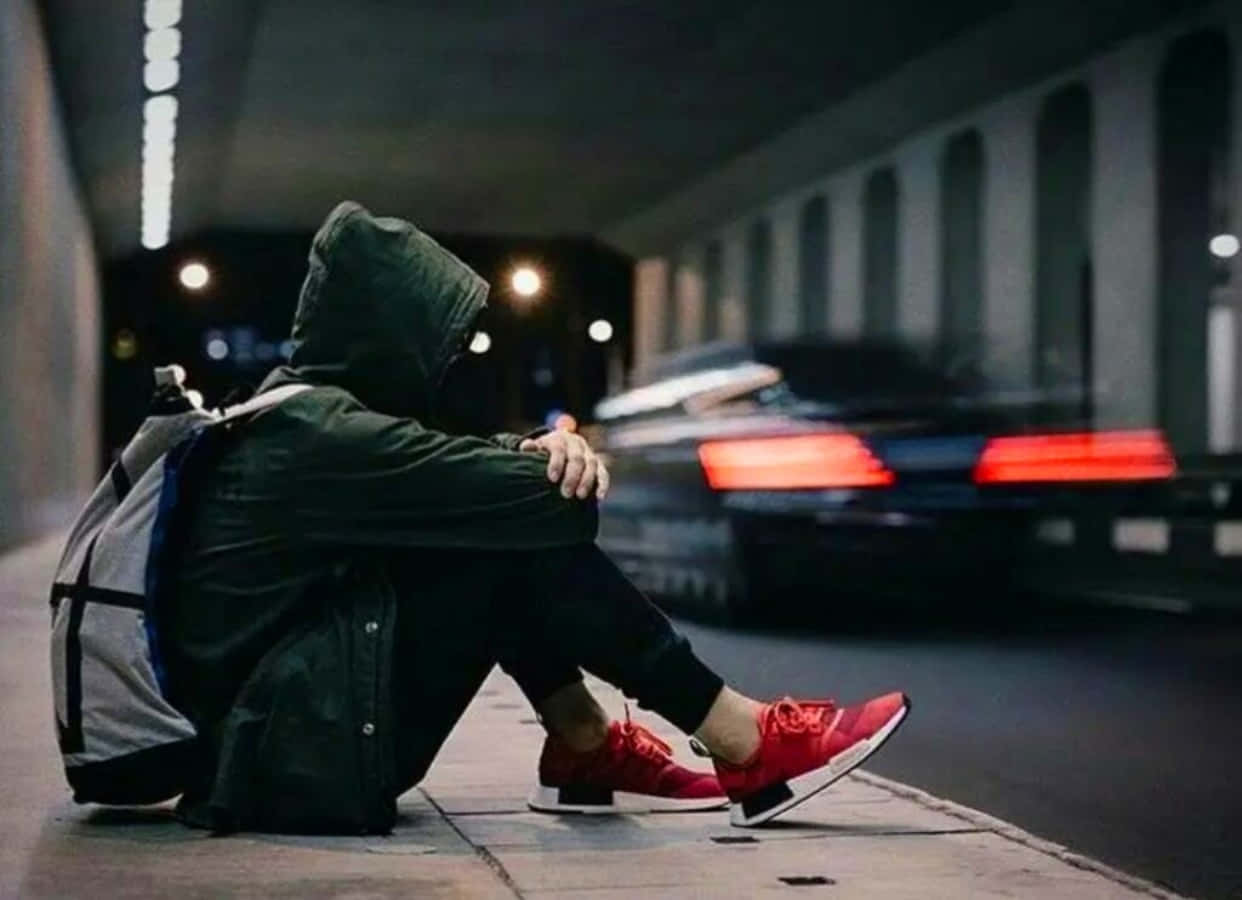 Alone 桌布 壁紙 — 一個戴著連帽衫的人獨自坐在夜晚的人行道上，一輛汽車帶著紅色尾燈疾馳而過。