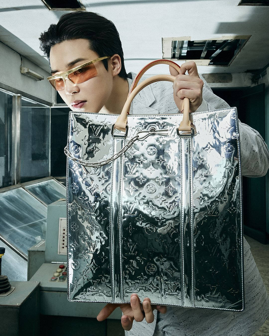 智旻 桌布 壁紙 — 一個戴著金色邊框太陽眼鏡的男人，在復古工業環境中拿著一個帶有壓花字母組合的大型銀色路易威登手提包。