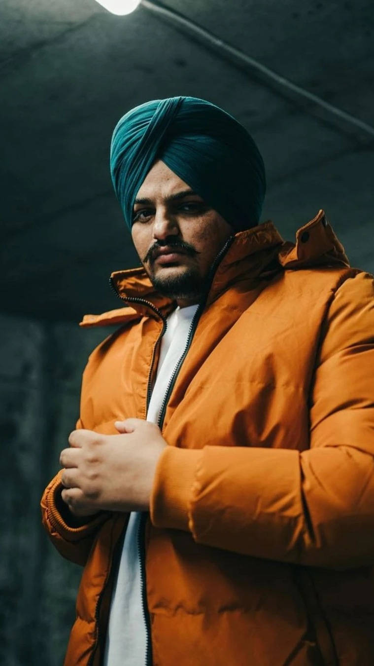 男人在昏暗的工業環境中戴著青綠色頭巾和橙色外套。 — Sidhu Moose Wala 壁紙