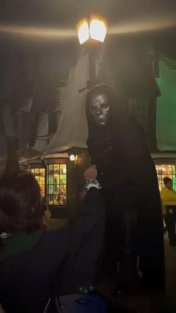Death Eaters 桌布 壁紙 — 一個戴著面具、身穿深色長袍的人物，在昏暗的主題景點中，於發光的街燈下籠罩著一位訪客。